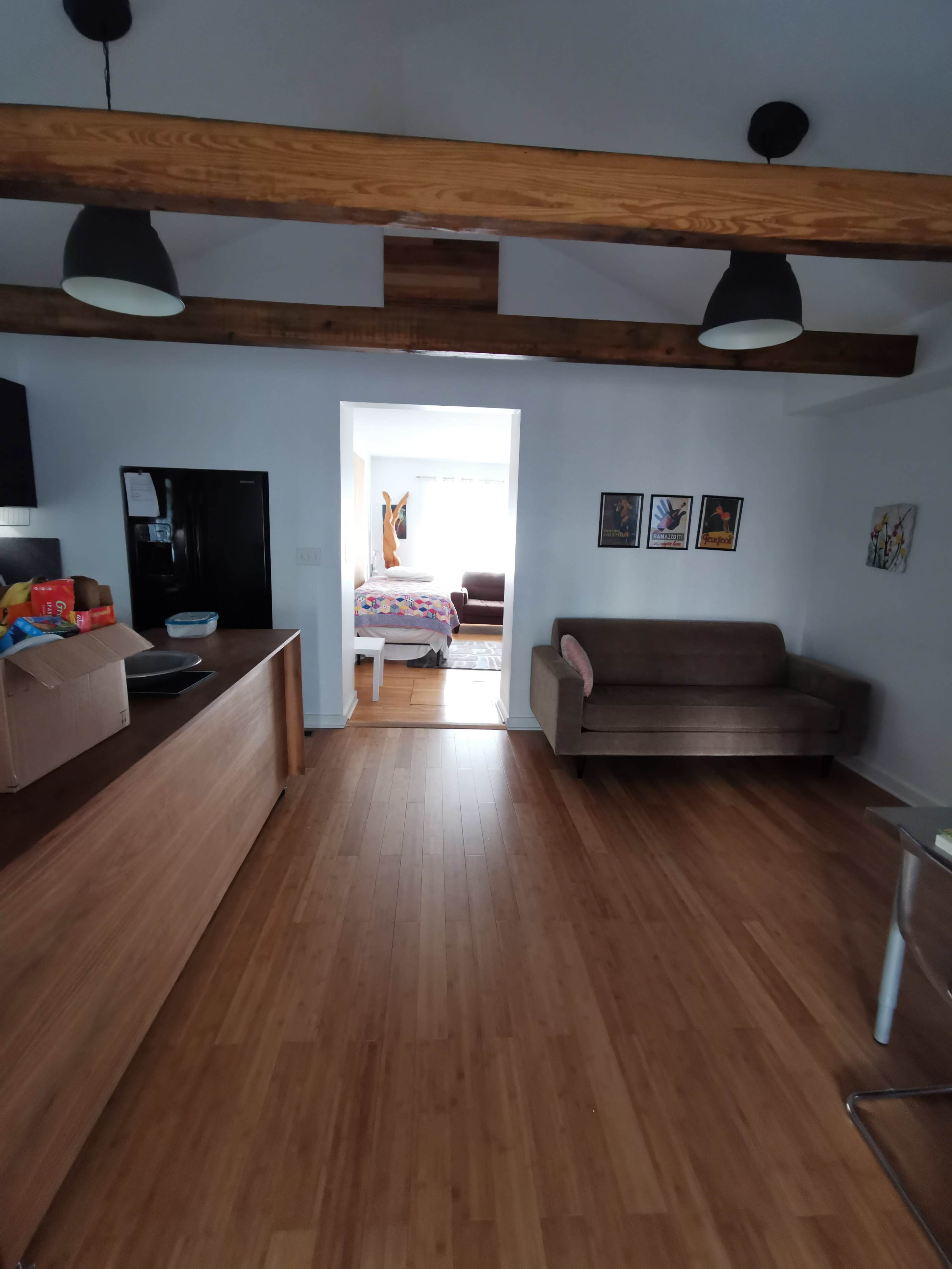A spacious interior with wooden beams, a kitchen area with a countertop, a brown sofa, and an open doorway leading to another room.