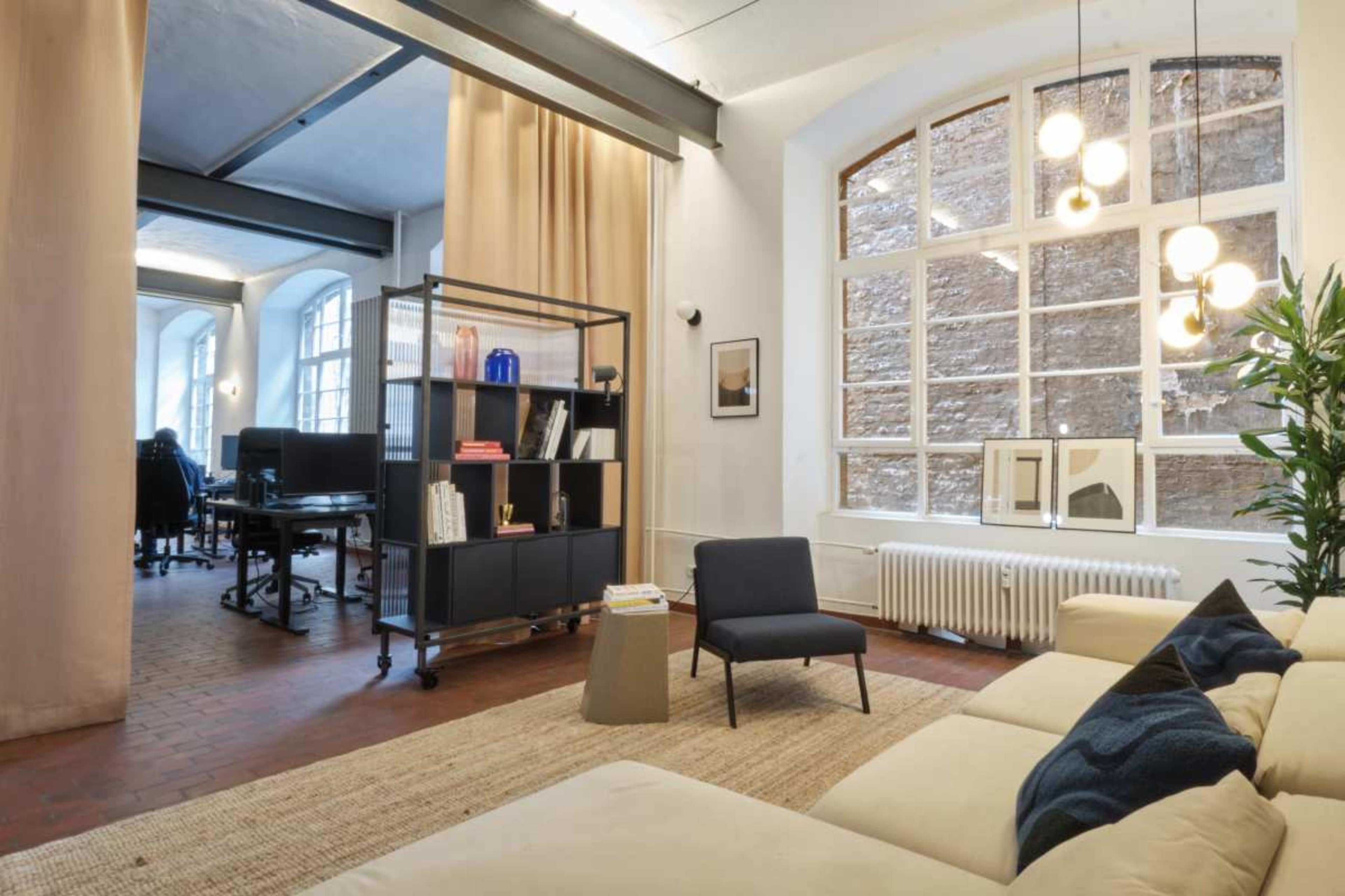 A spacious office interior features a seating area with a light-colored sofa and a shelving unit, while a work area is visible in the background.