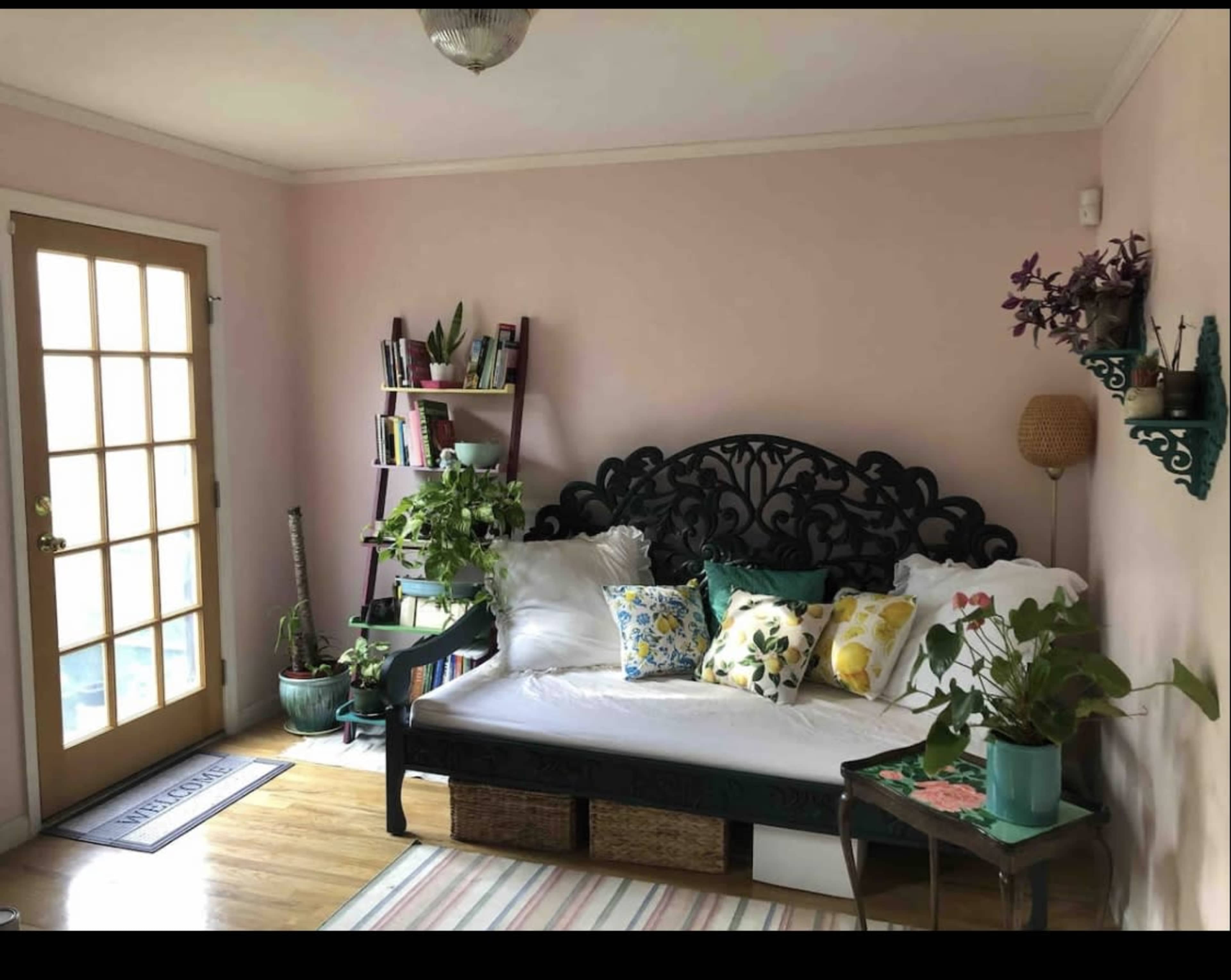 The room features a daybed adorned with decorative pillows, a tall bookshelf filled with plants and books, and natural light streaming through a glass door.
