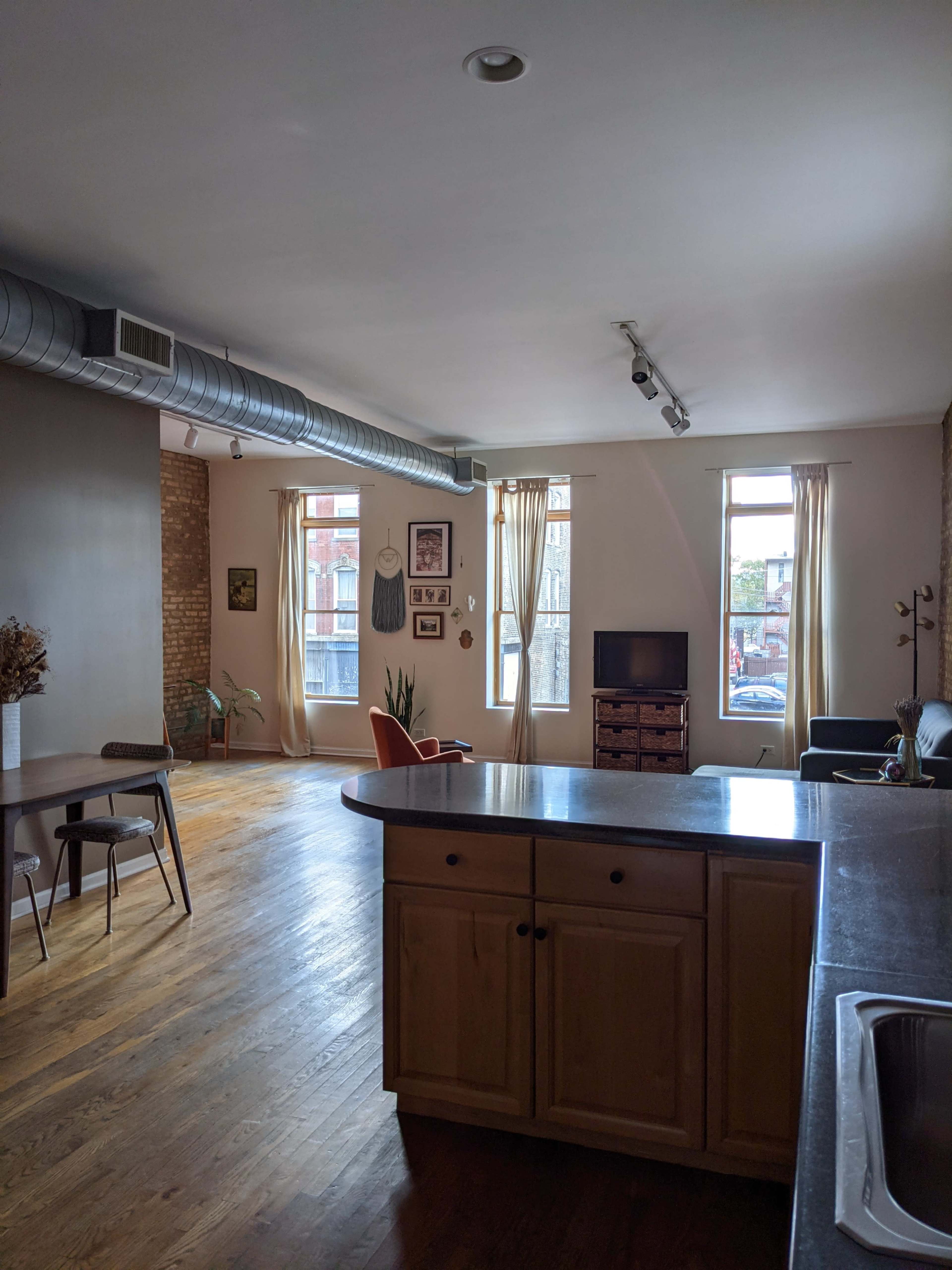 The image shows a spacious living area with a kitchen island, featuring wood flooring, large windows, and minimal furniture, including an orange chair and a television.