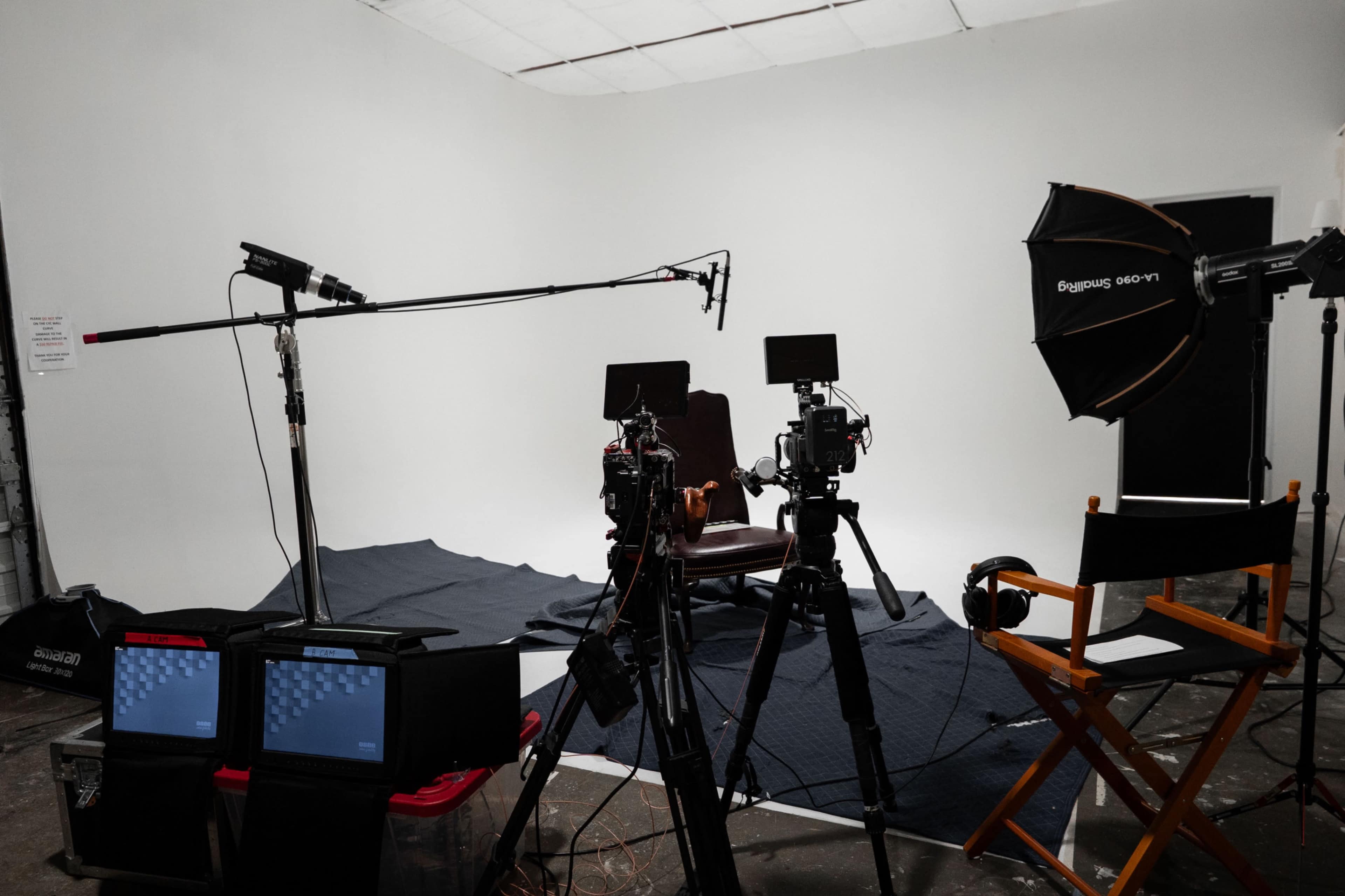 The image shows a film or photography studio setup with a wooden chair, multiple cameras, lighting equipment, and a sound recording microphone against a plain white backdrop.