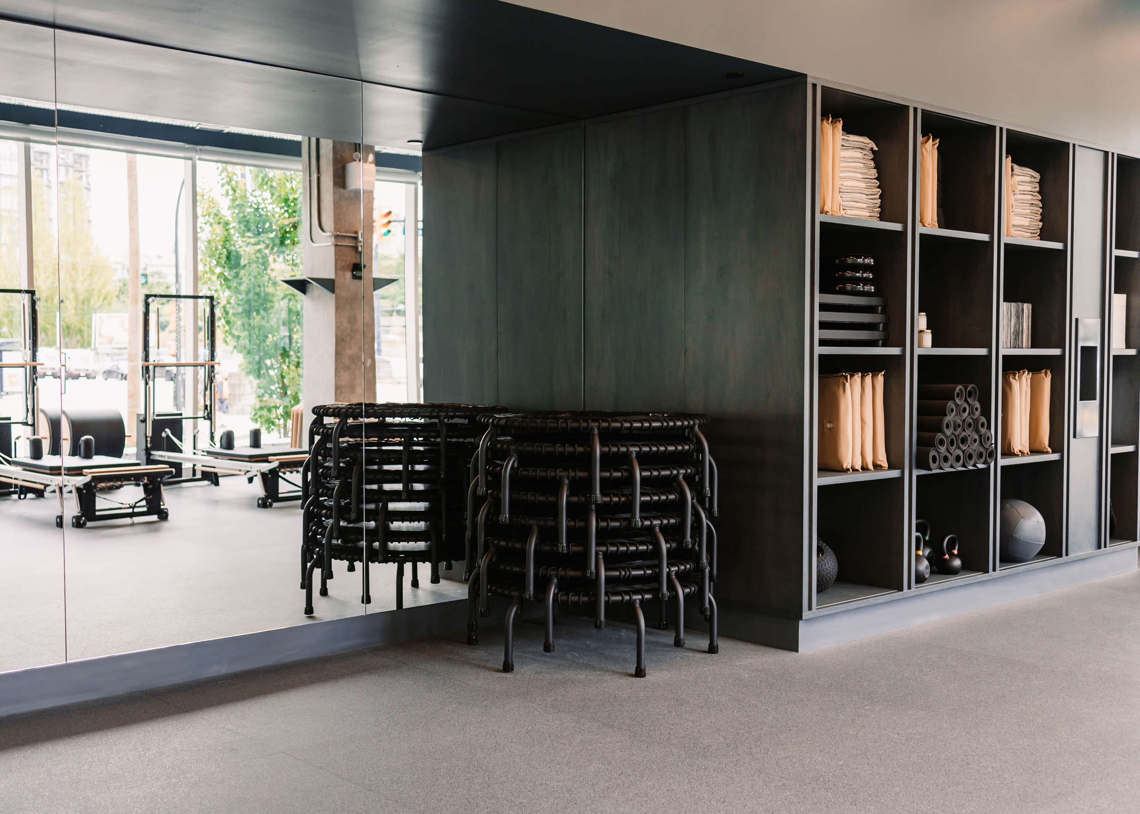 The image shows a modern fitness studio with neatly stacked black mats and organized storage shelves filled with exercise equipment and rolled yoga mats.