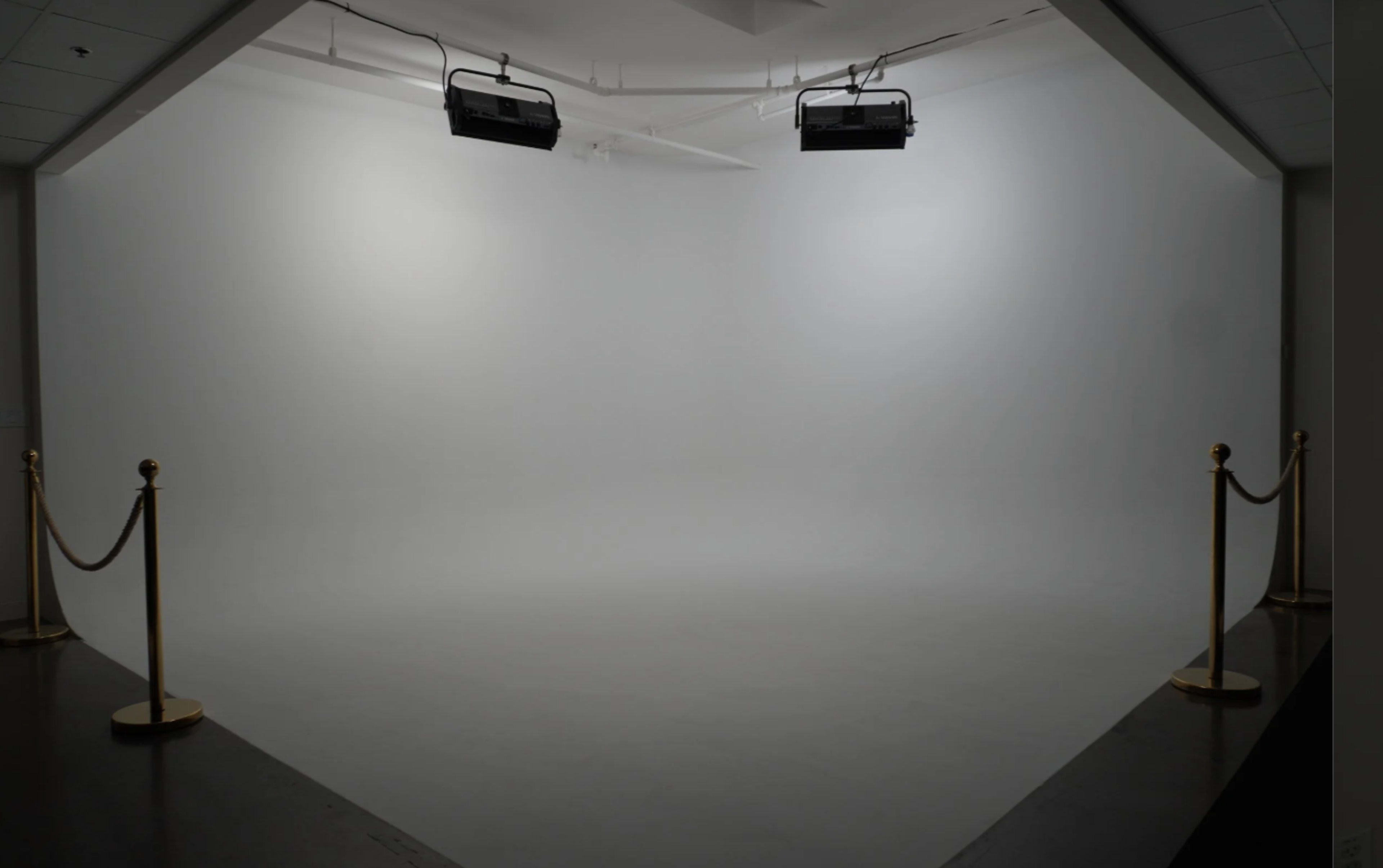The image shows a blank white studio space with two overhead lights and a velvet rope barrier.