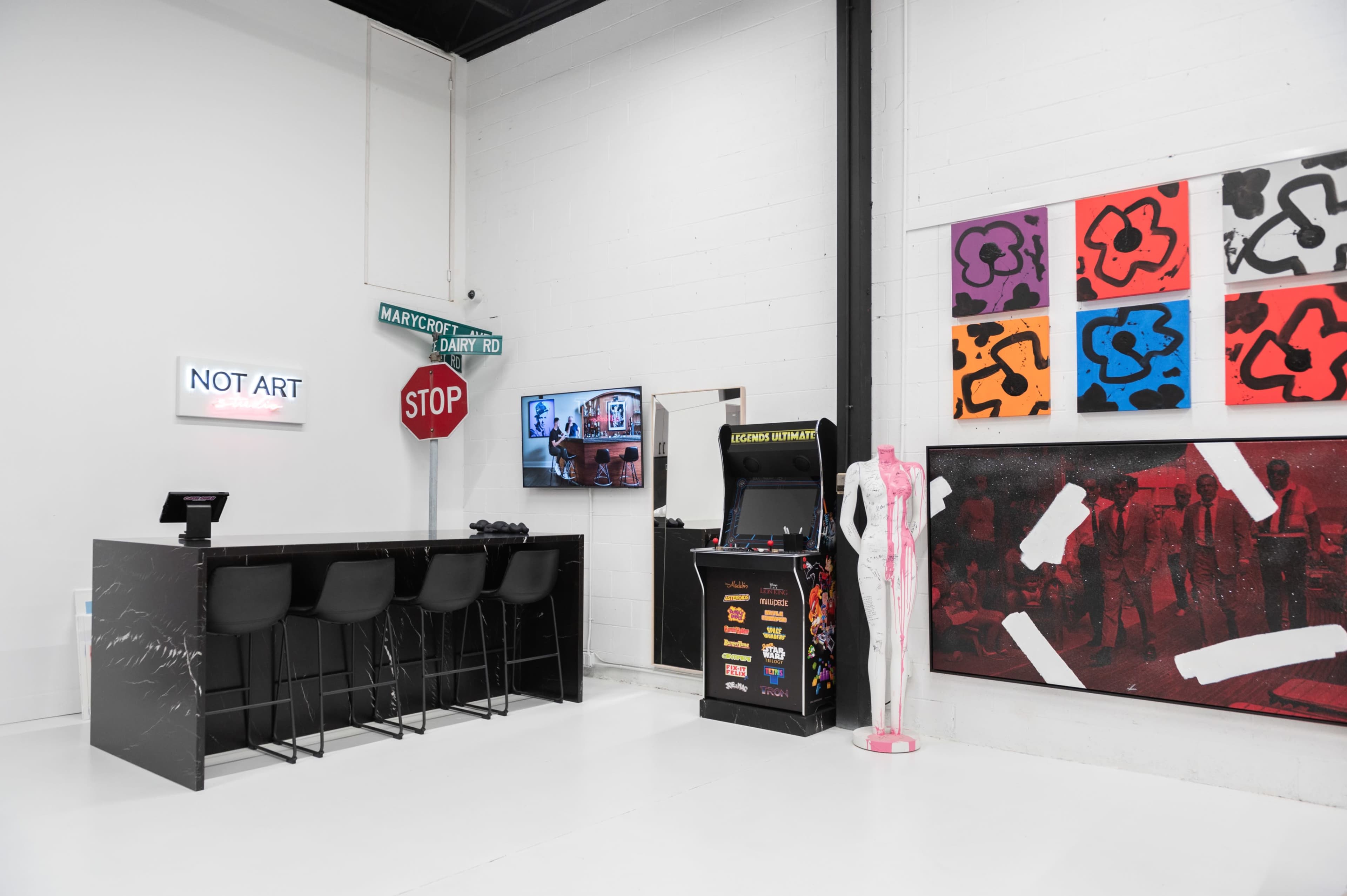 A modern gallery space with a bar area featuring stools, a stop sign, an arcade machine, and colorful artwork on the walls.