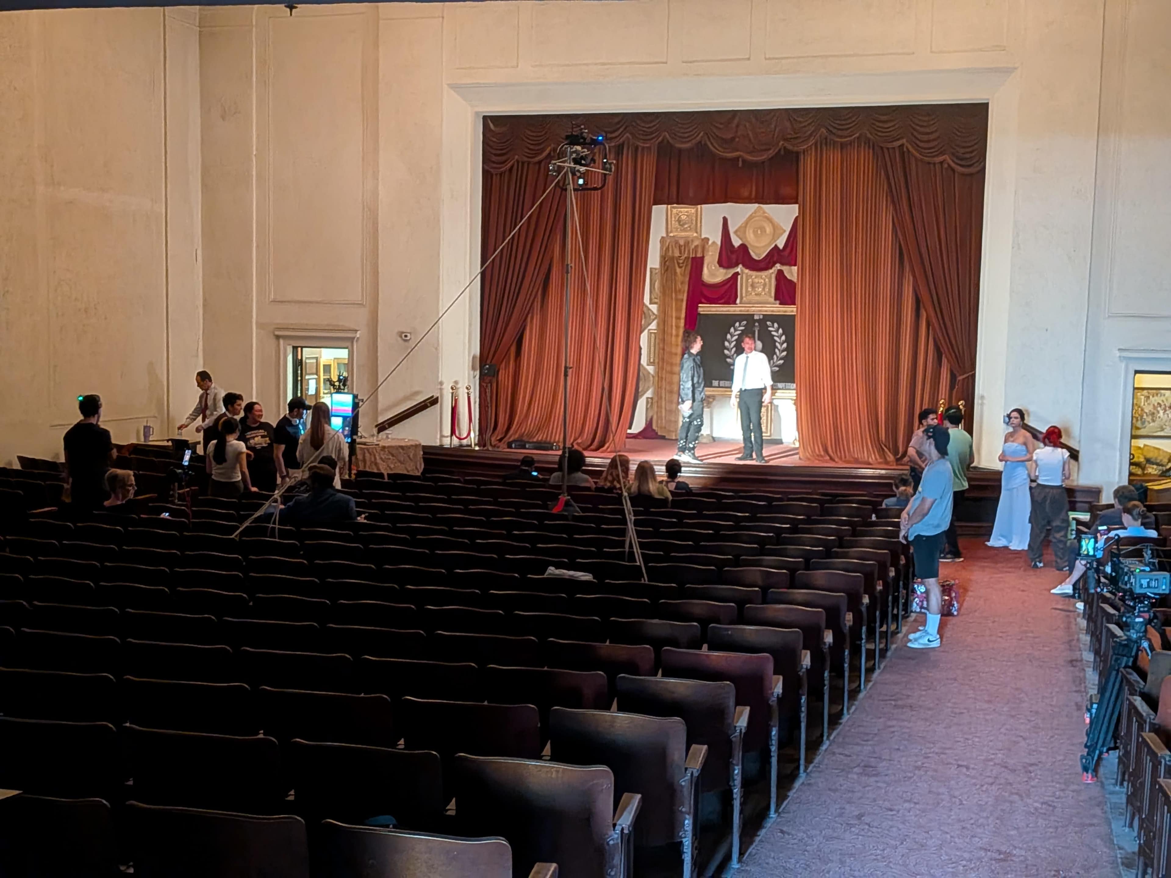 A film crew sets up in an empty theater with a large curtain backdrop and numerous people scattered throughout the seating area.