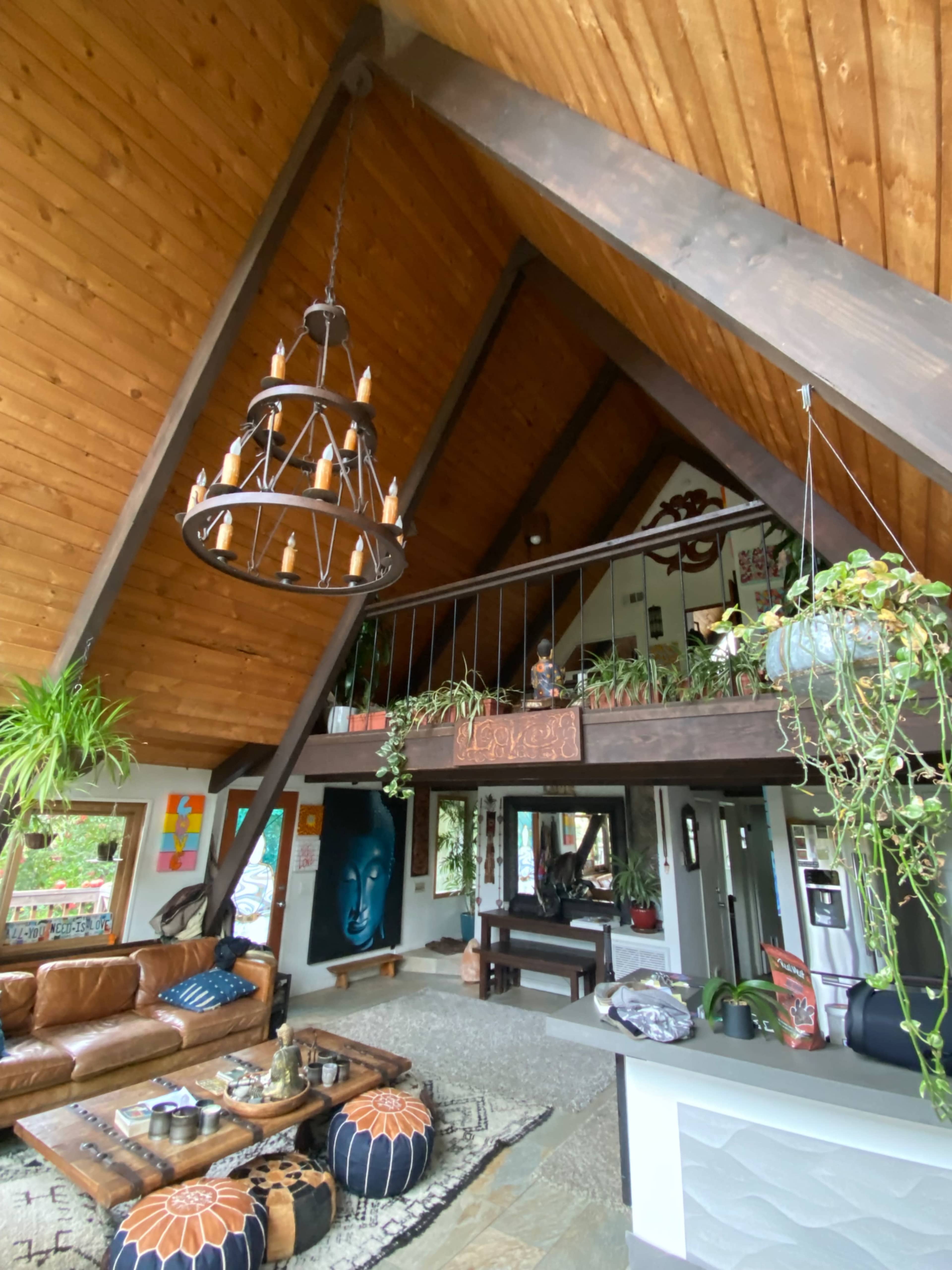 An interior of a spacious, A-frame living area with a wooden ceiling, a balcony, and various plants, featuring a leather sofa and a patterned rug.