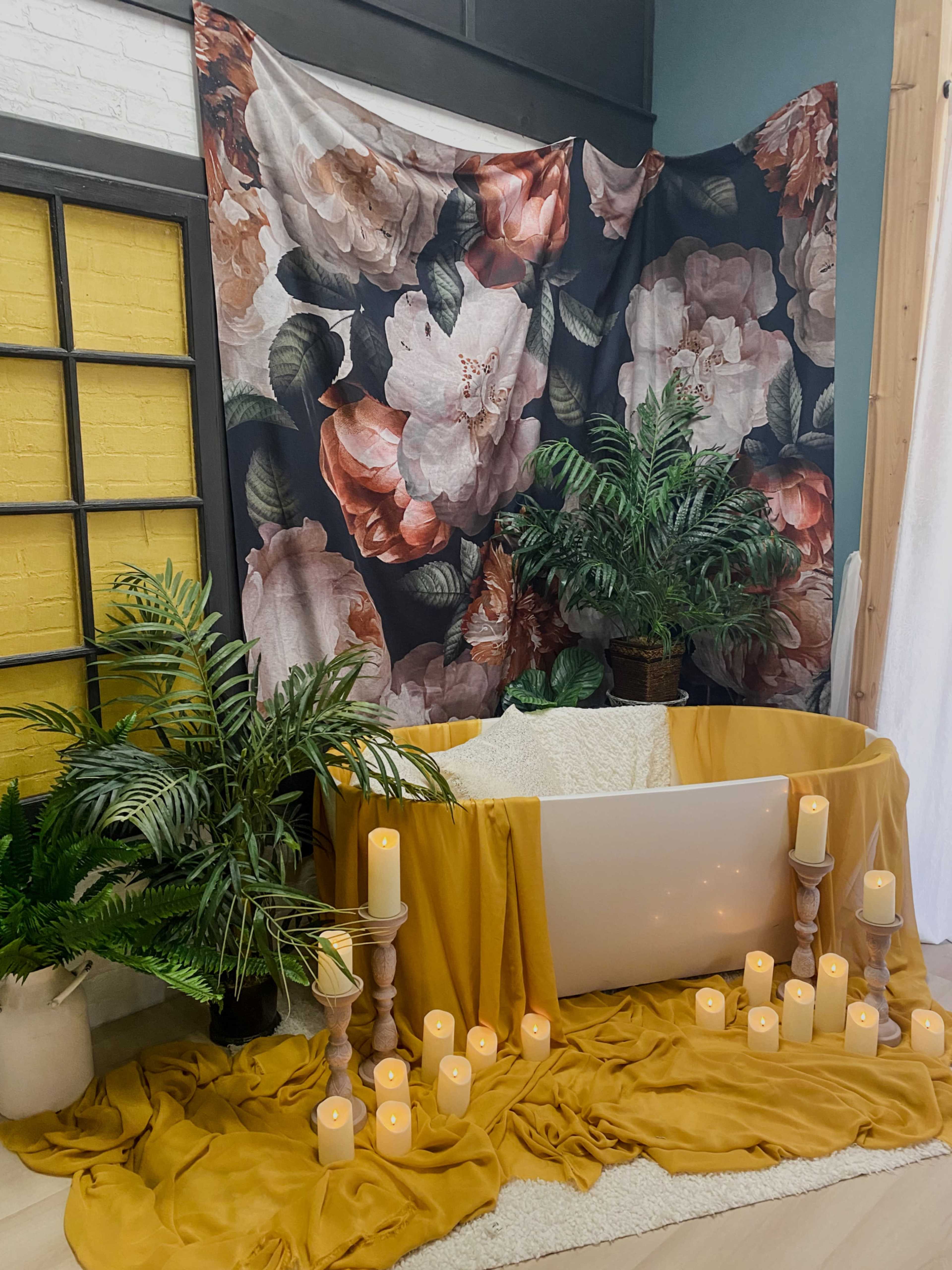 The image features a freestanding white bathtub surrounded by draped fabric, decorative candles, and potted plants, set against a floral backdrop.