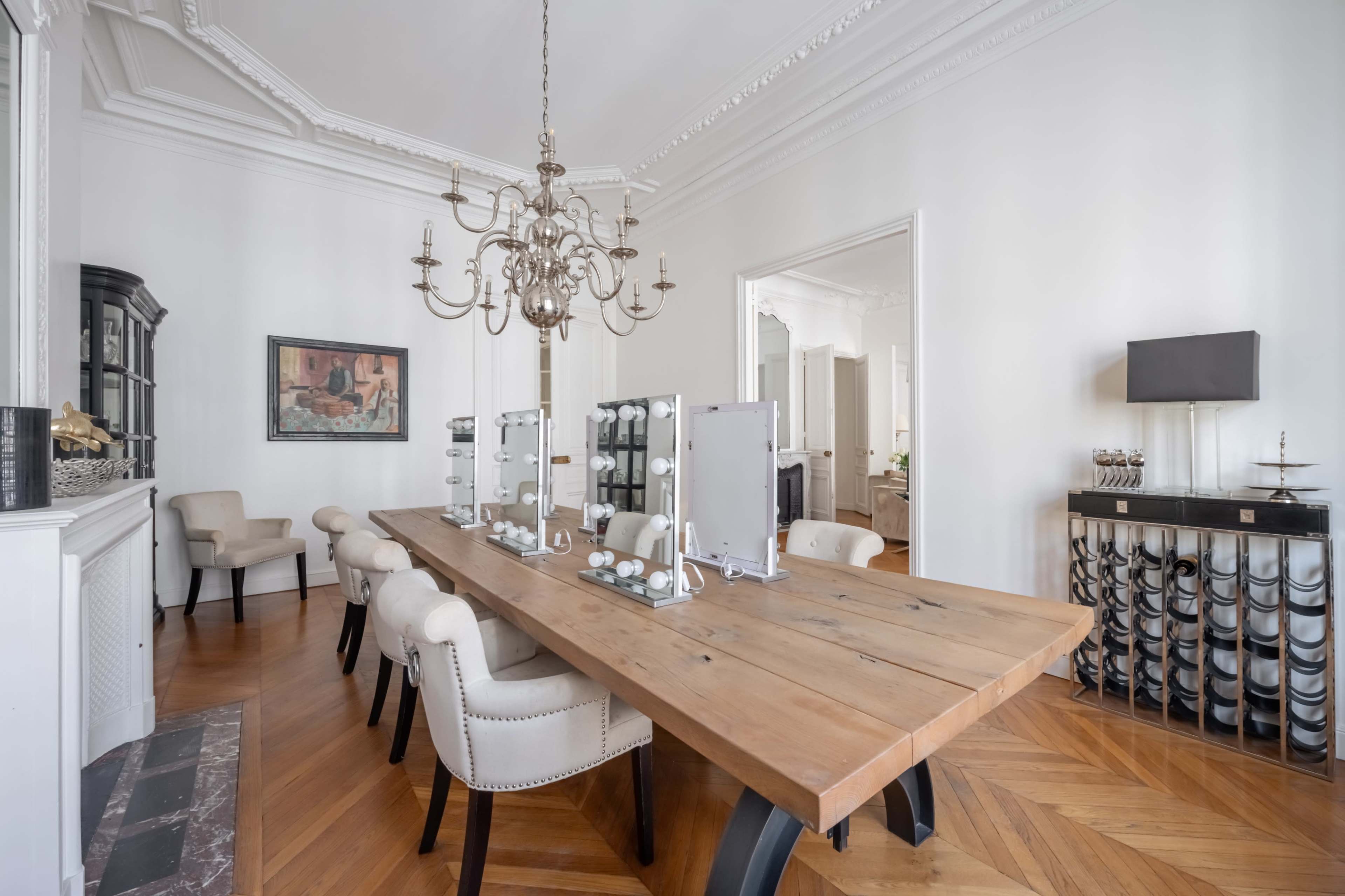 The image shows a spacious dining room featuring a wooden table with chairs, a chandelier, and mirrors along one side.