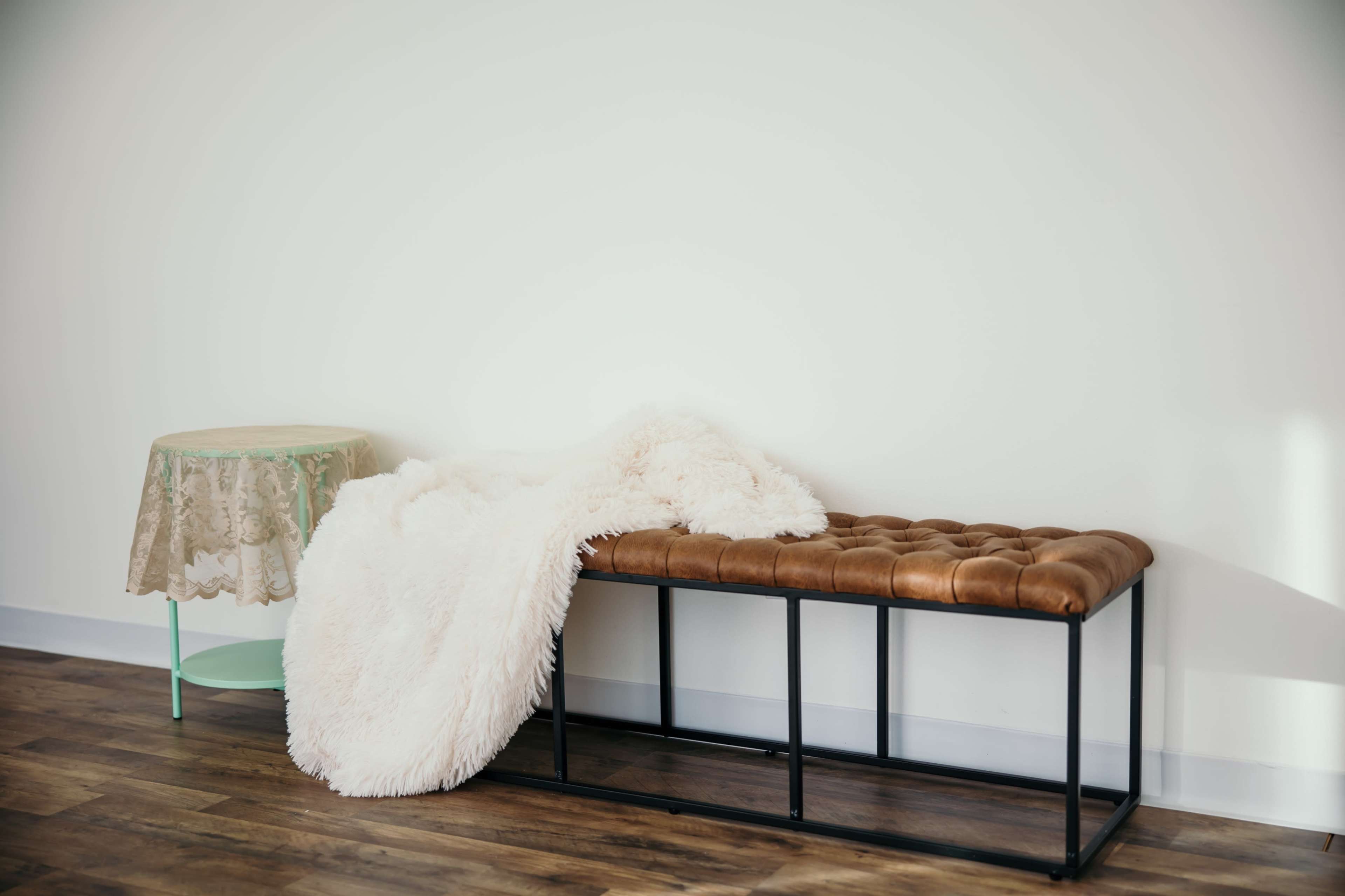 A cozy space features a leather bench with a fluffy throw draped over it beside a circular table covered with a floral tablecloth.