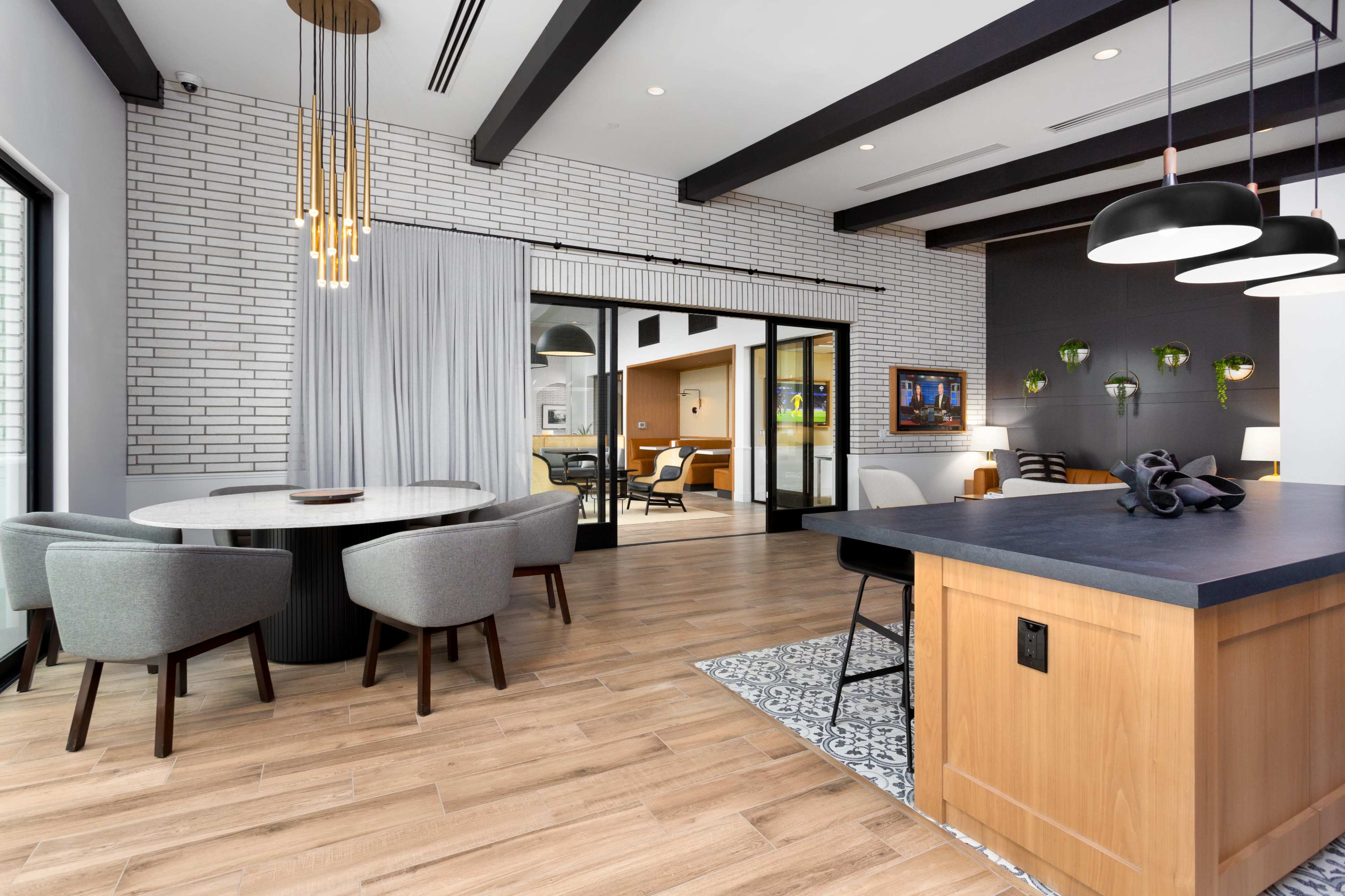 A modern open-concept dining and living area featuring a round table with gray chairs, a kitchen island, and large windows with a brick accent wall.