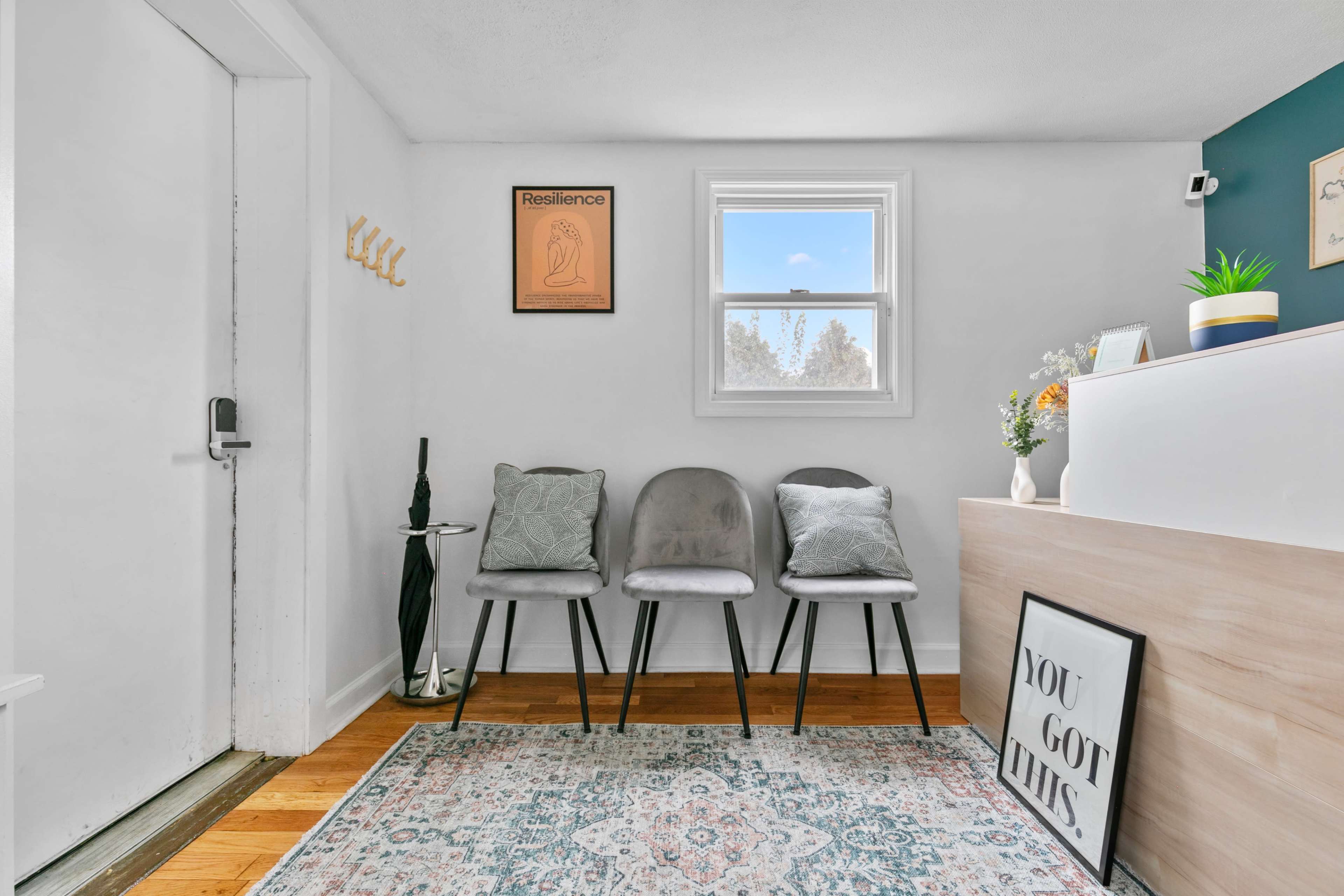 A small entryway features three gray chairs, a light wood reception desk, and a window allowing natural light to enter, with a decorative plant and artwork on the walls.