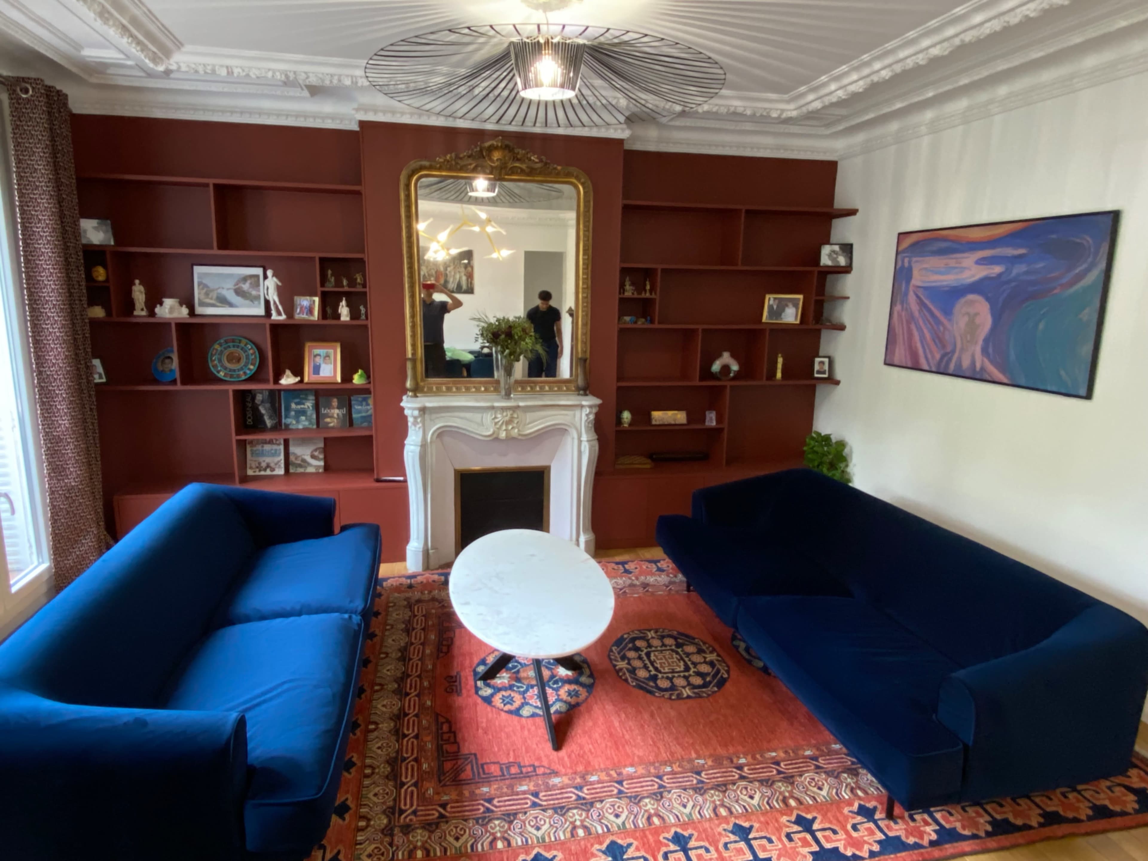 The image shows a living room with two blue sofas facing a white marble fireplace, a circular coffee table at the center, and a bookshelf along the back wall filled with various items and decorations.