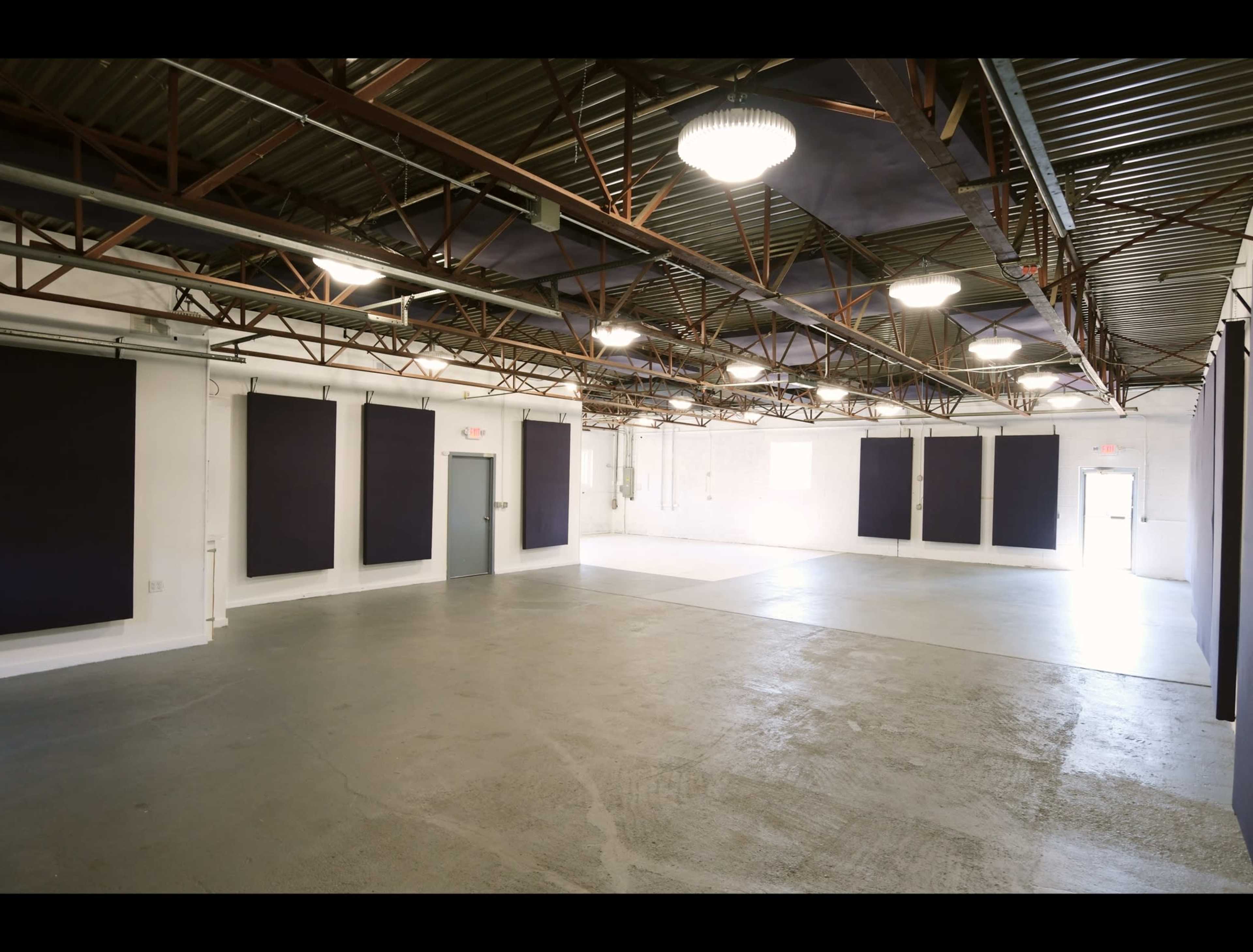 The image shows a spacious interior of an empty hall with high ceilings, concrete flooring, and dark curtains on the walls.