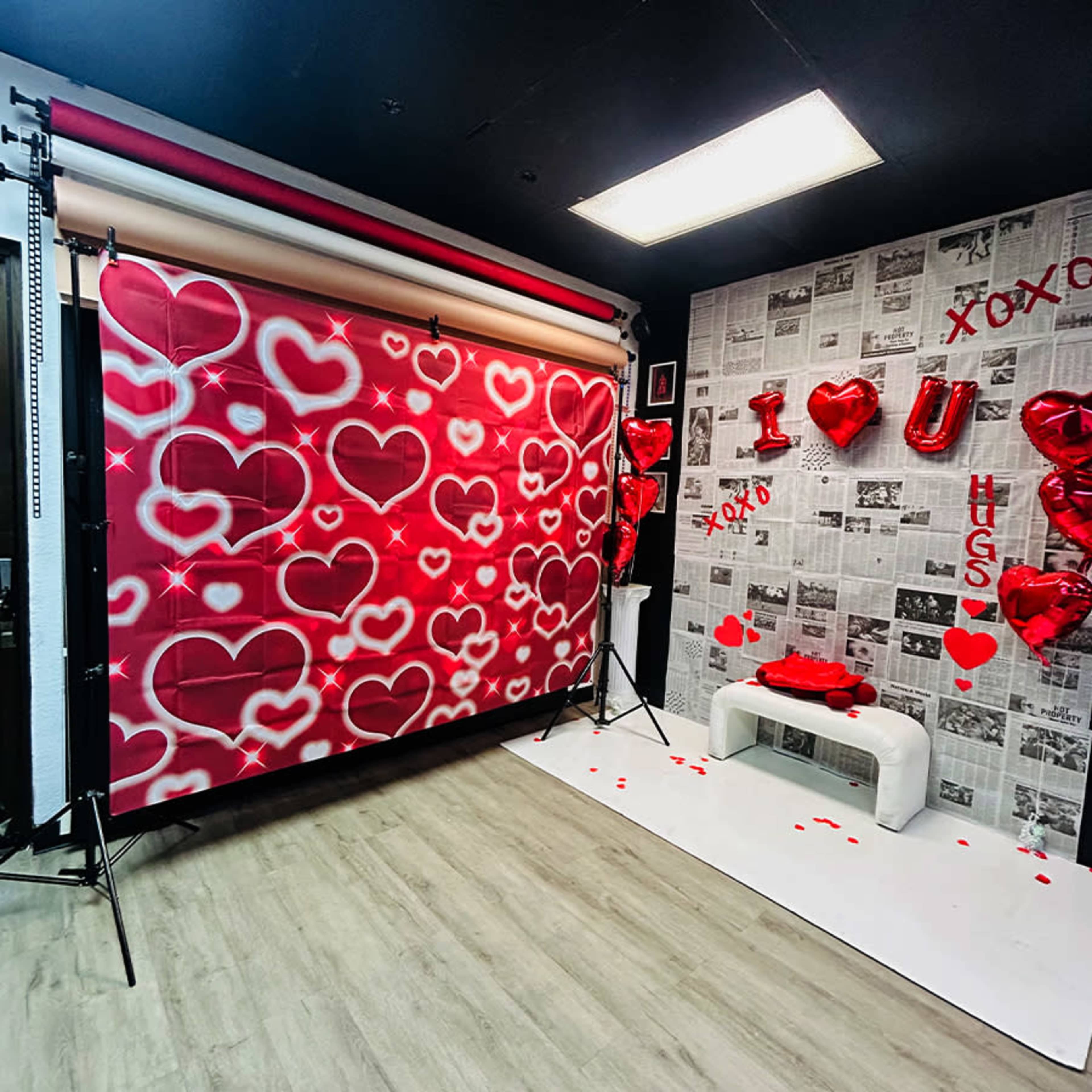The image features a photography backdrop with a red heart pattern, surrounded by decorated walls and floor with heart-shaped balloons and newspaper clippings.