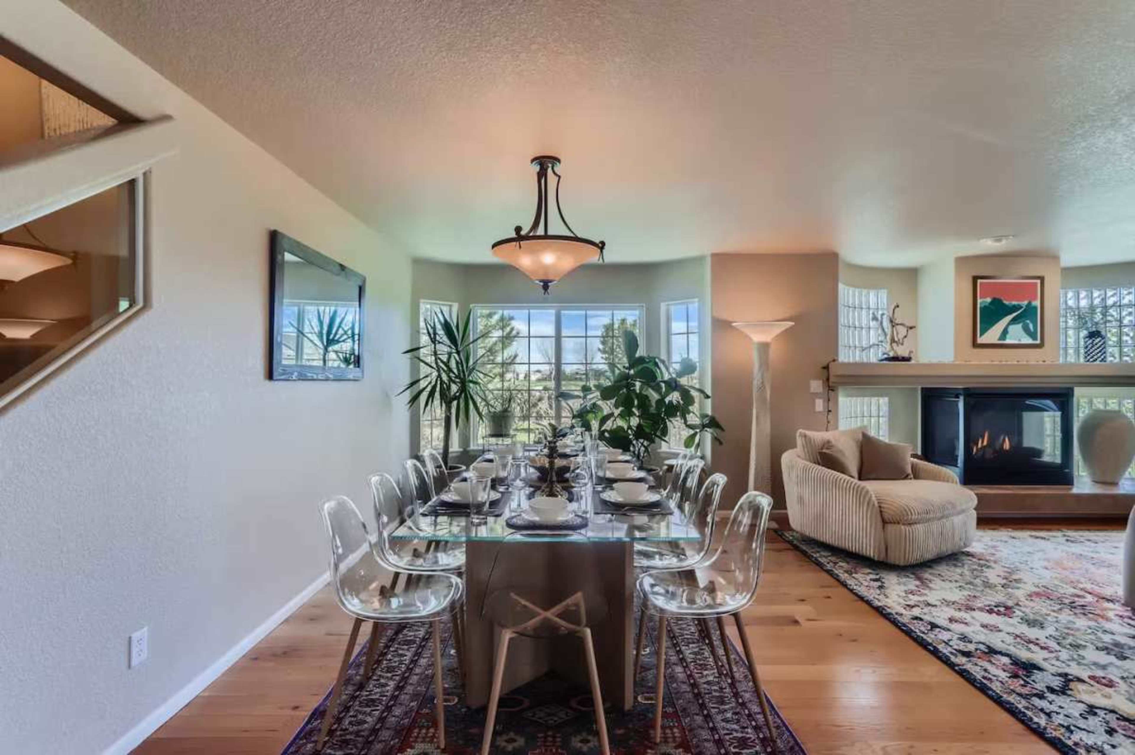 A long dining table set for a meal is positioned near large windows, with a cozy fireplace and plants in the background.