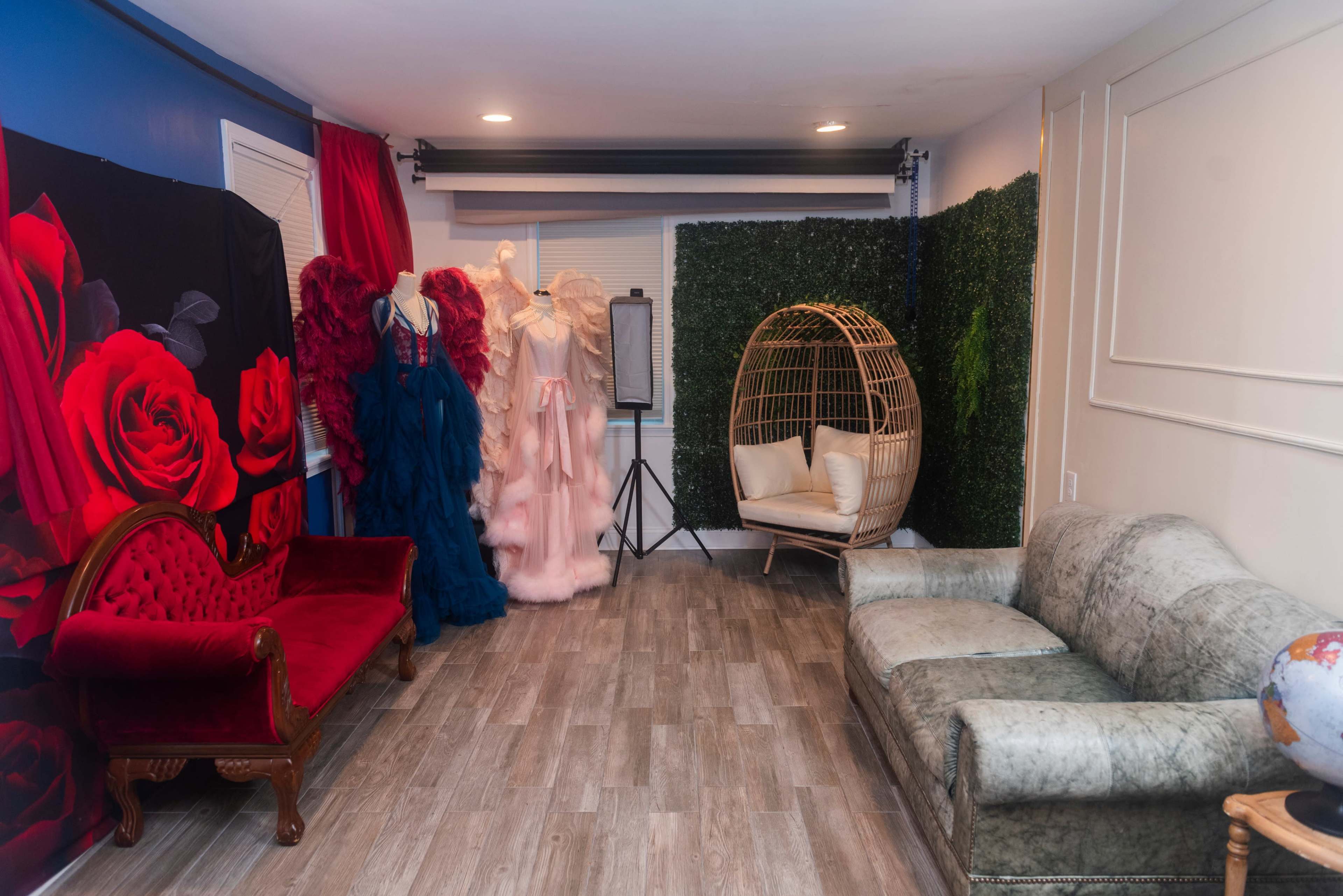 The scene depicts a styled room featuring a red velvet couch, a grey upholstered sofa, a clothing rack with elegant dresses, and a wicker chair surrounded by artificial greenery.