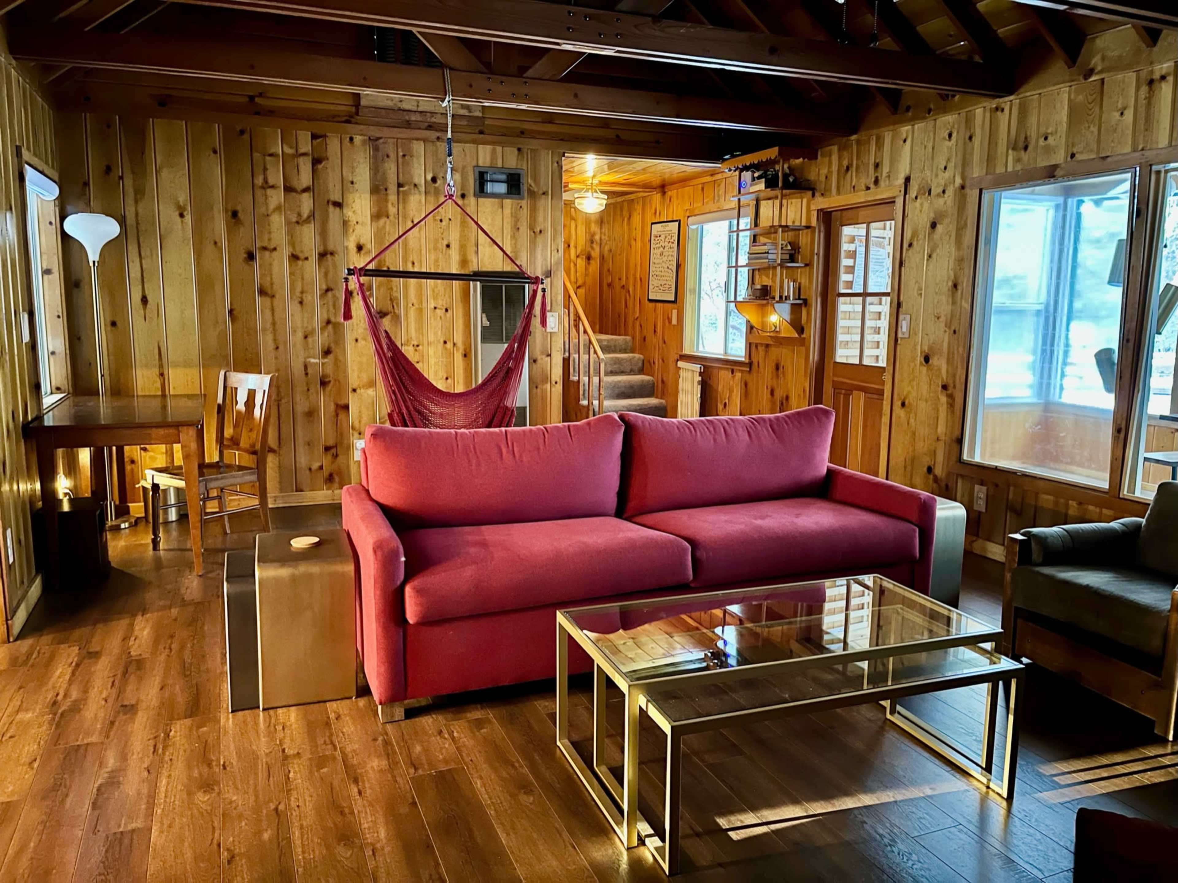 A wooden cabin interior featuring a red sofa, a glass coffee table, a hammock, a dining area, and large windows letting in natural light.