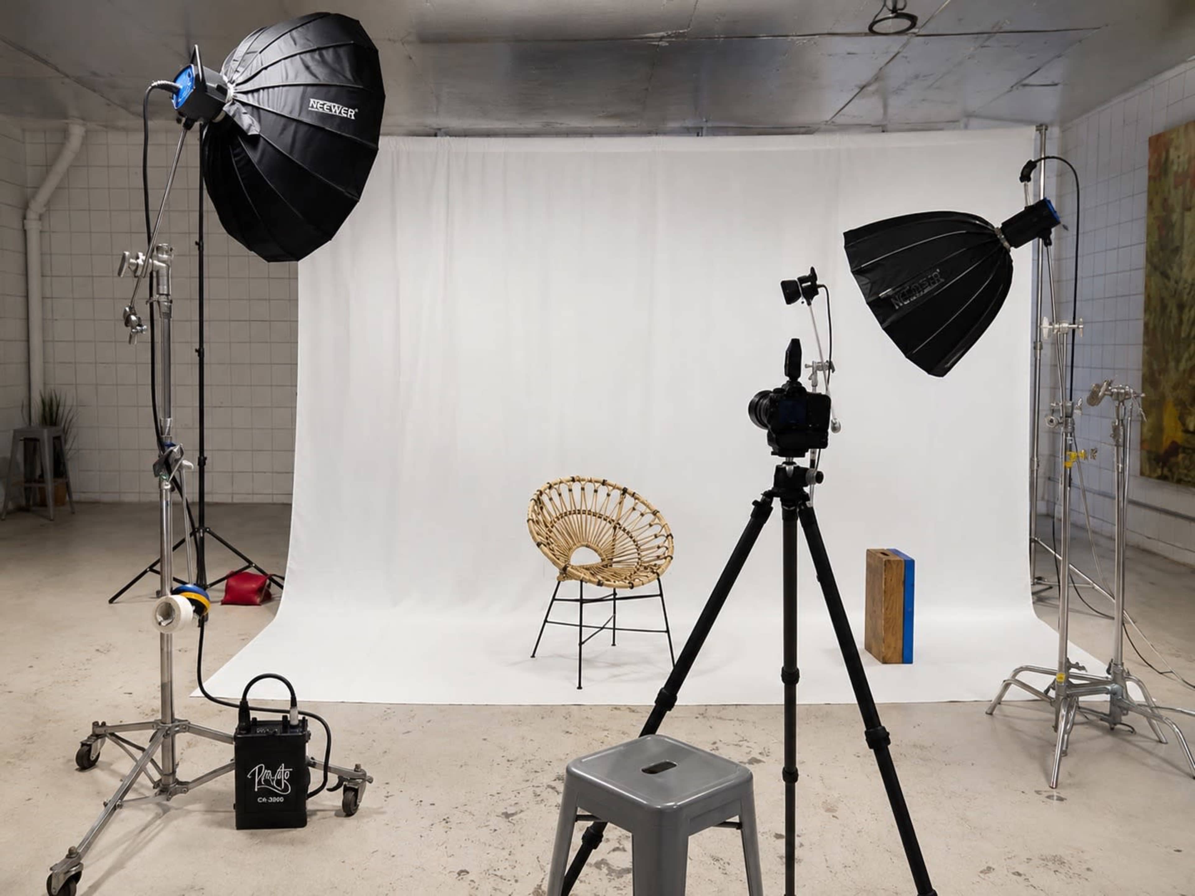 The image shows a photography studio setup with two light stands, a camera on a tripod, a rattan chair, and a backdrop of white fabric.