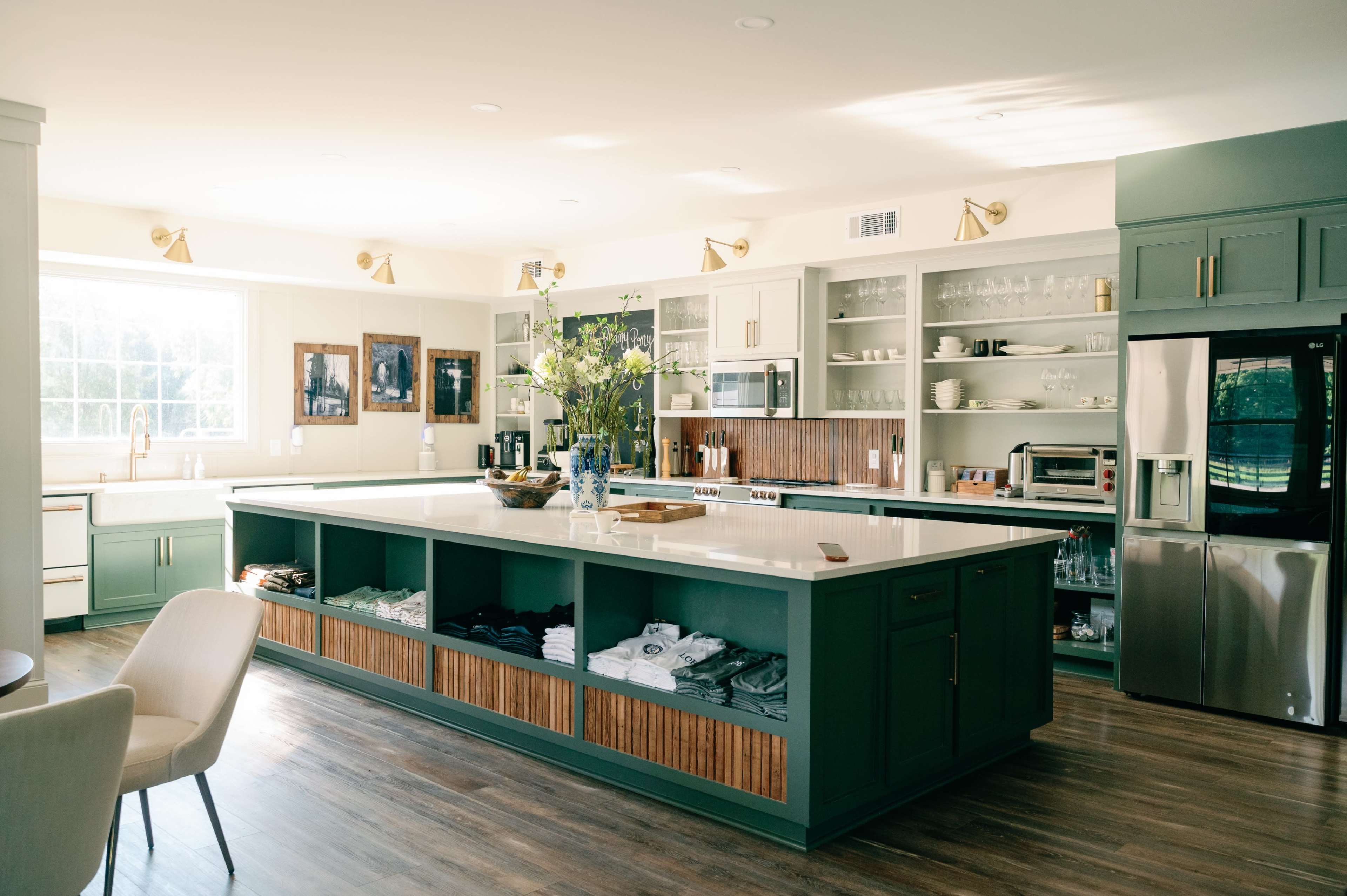 The image shows a modern kitchen with a large island, green cabinetry, a sink under a window, and shelves displaying glassware and kitchen items.