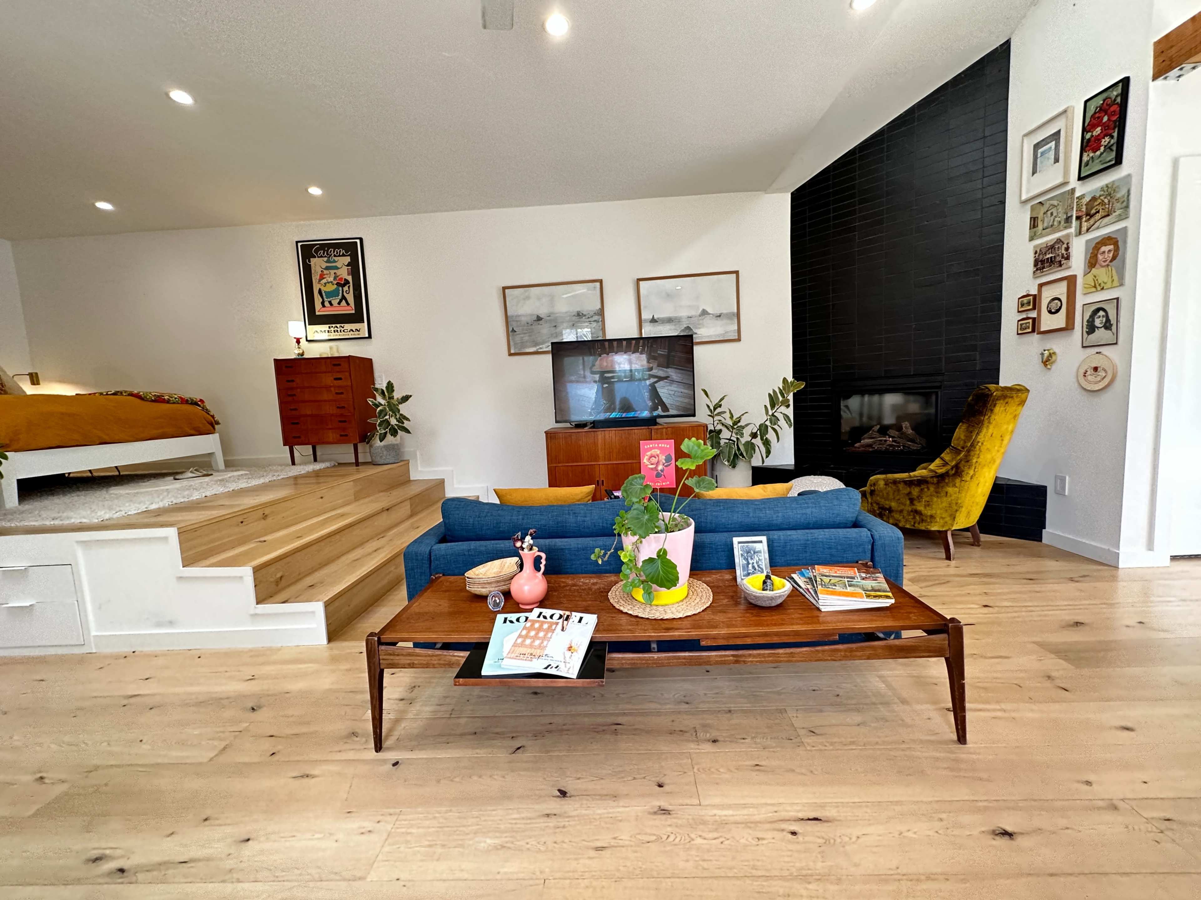 A cozy living room features a blue sofa, wooden coffee table with decorative items, and a television, adjacent to a staircase and a dining area.