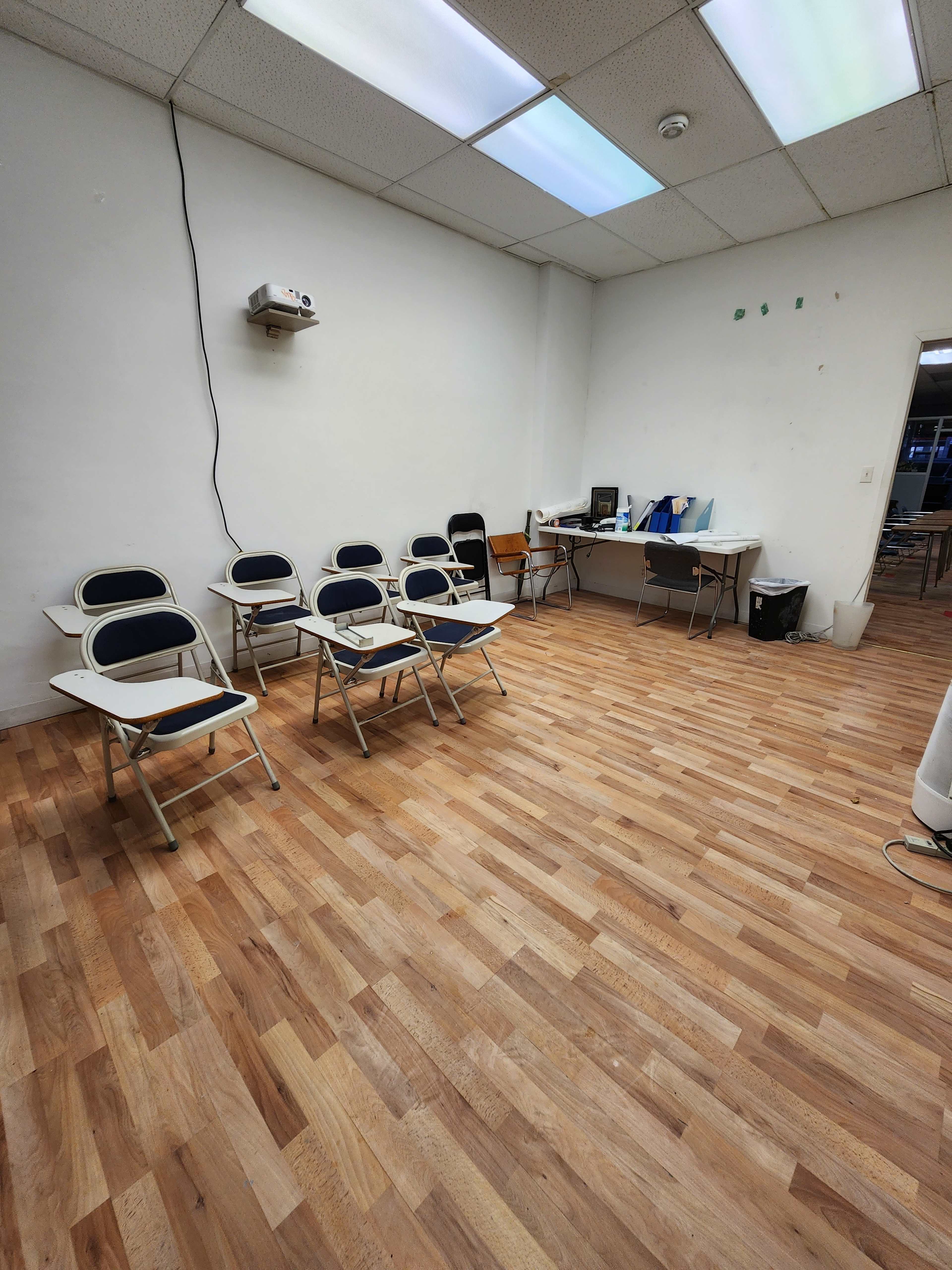 A sparsely furnished room with several chairs arranged in a row and a table in the corner, featuring a computer and other items.