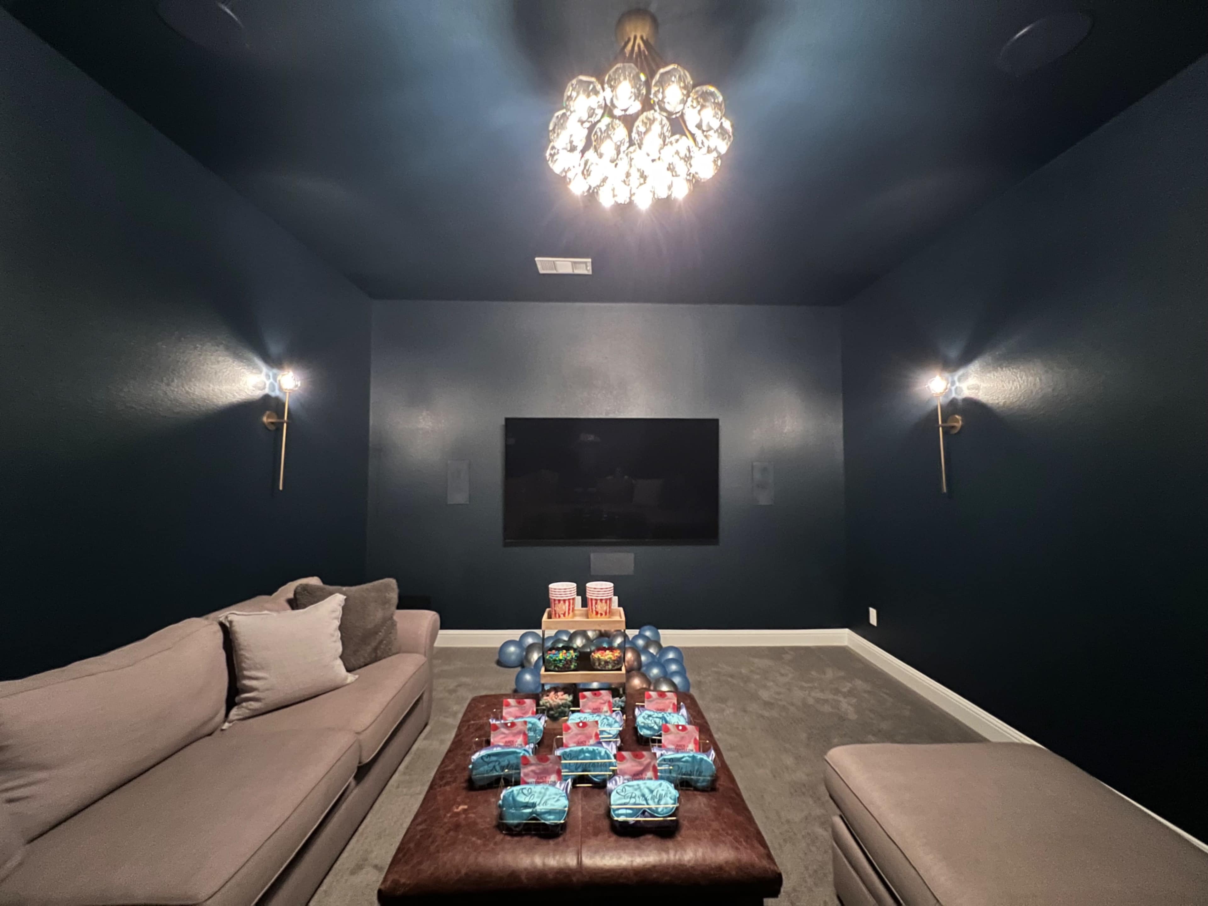A dimly lit home theater features a large TV on the wall, a plush sectional couch, and a coffee table adorned with colorful snacks.
