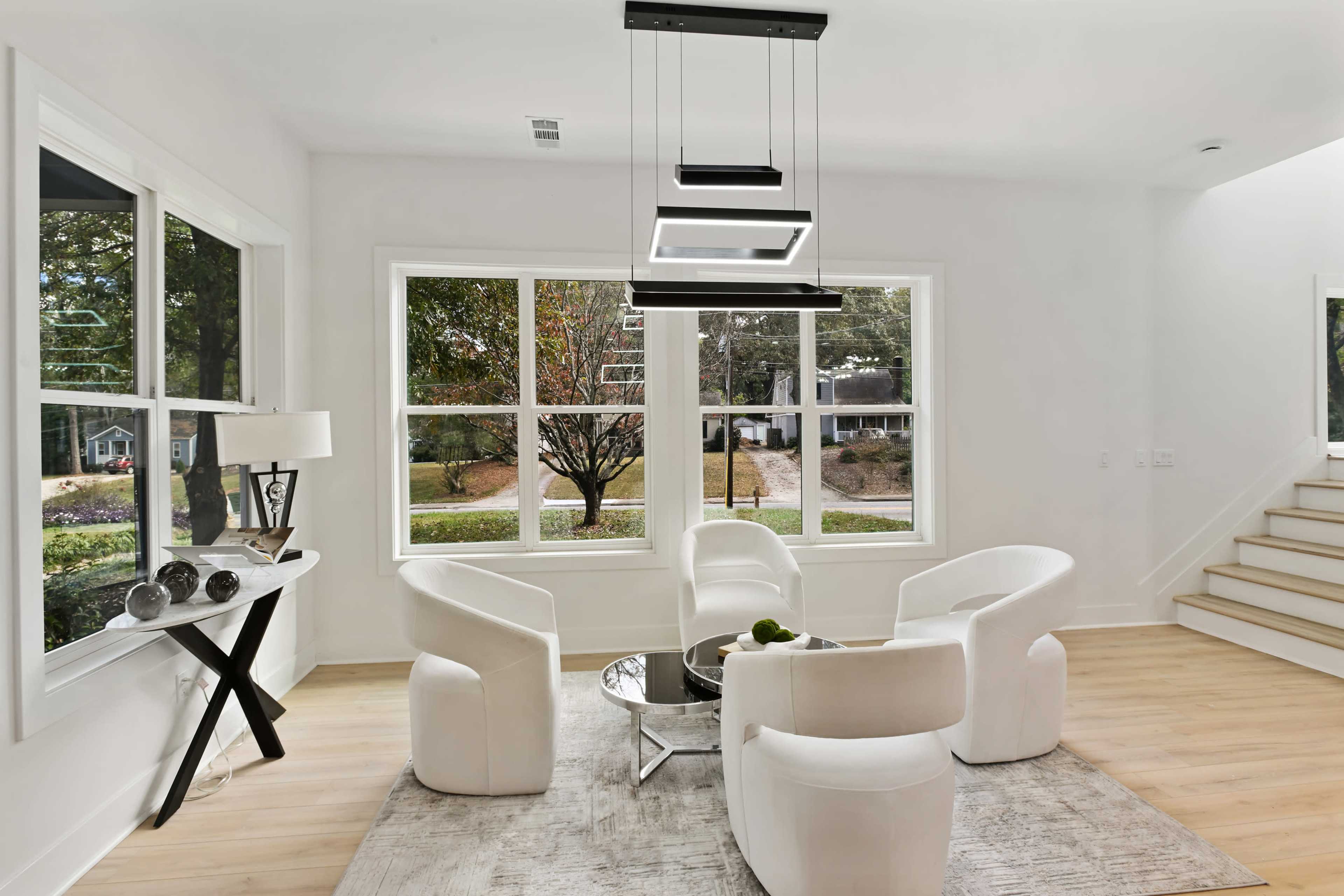 A modern living room features four white upholstered chairs around a circular coffee table, with large windows providing a view of the outdoor landscape.