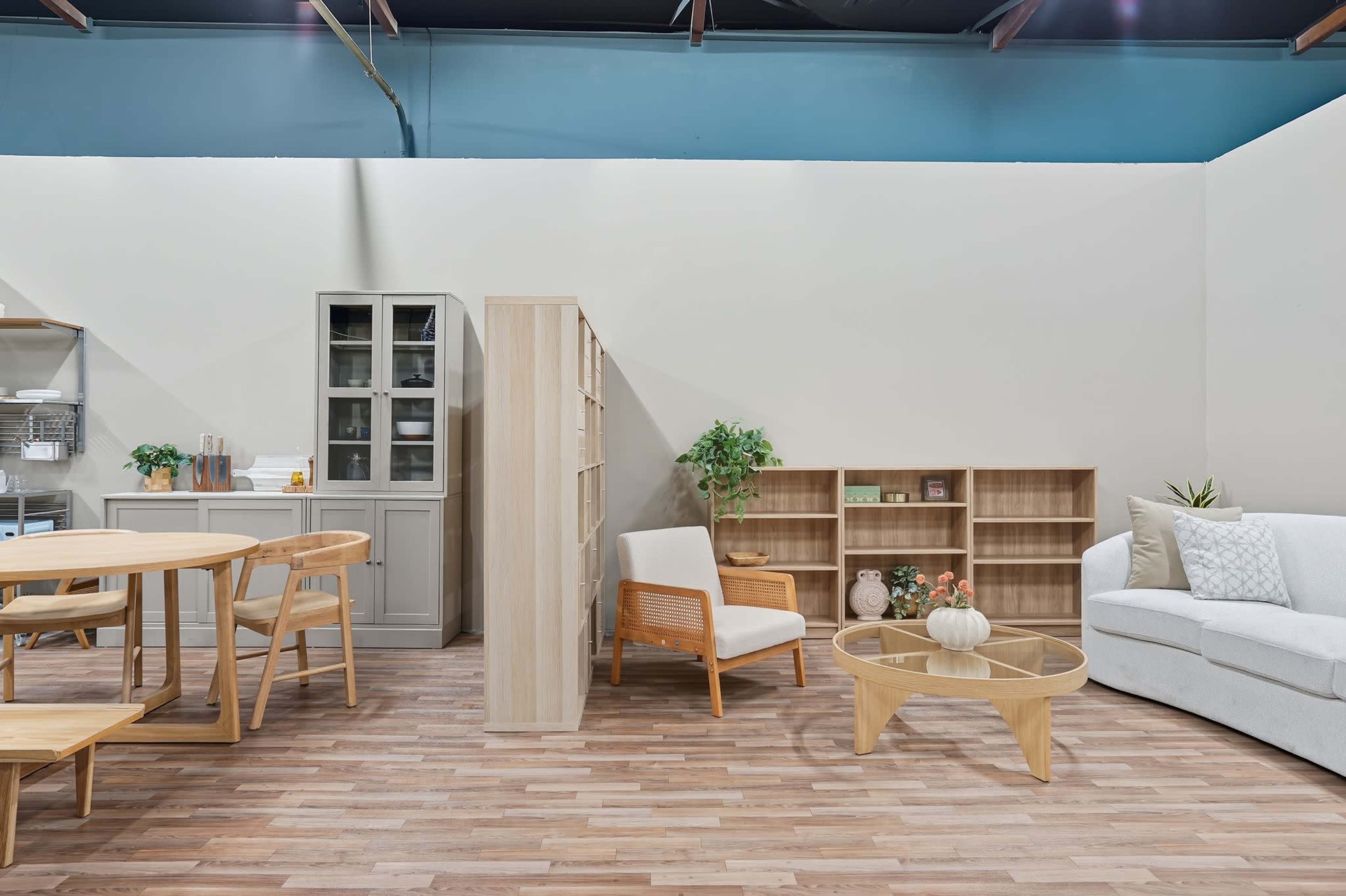 The image shows a spacious interior design display featuring a light wooden dining table and chairs, a modern white sofa, and a combination of shelving units with decorative items against a neutral wall.
