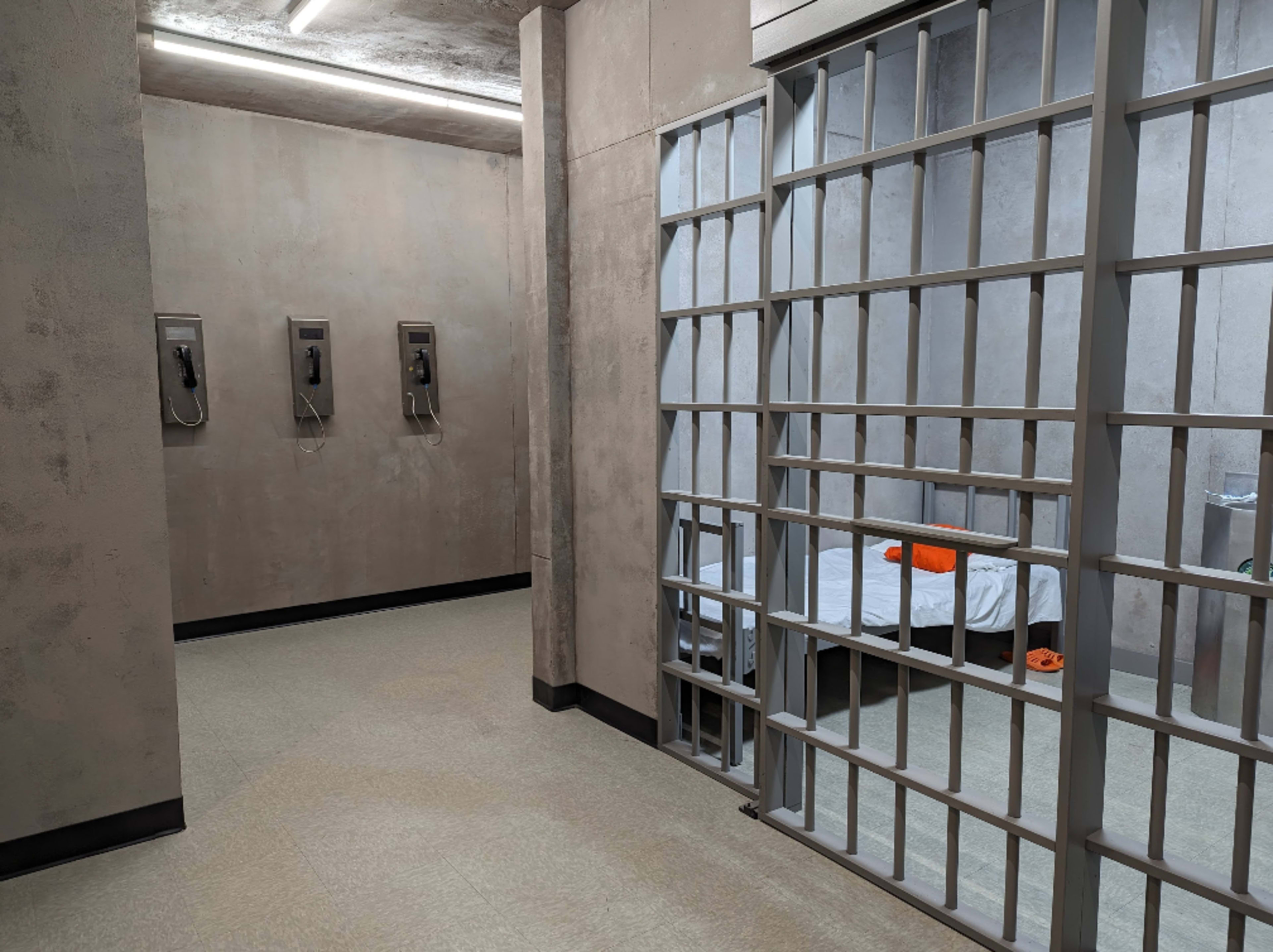 The scene shows the interior of a prison cellblock with a barred cell, a cot with bedding, and phone booths along the wall.