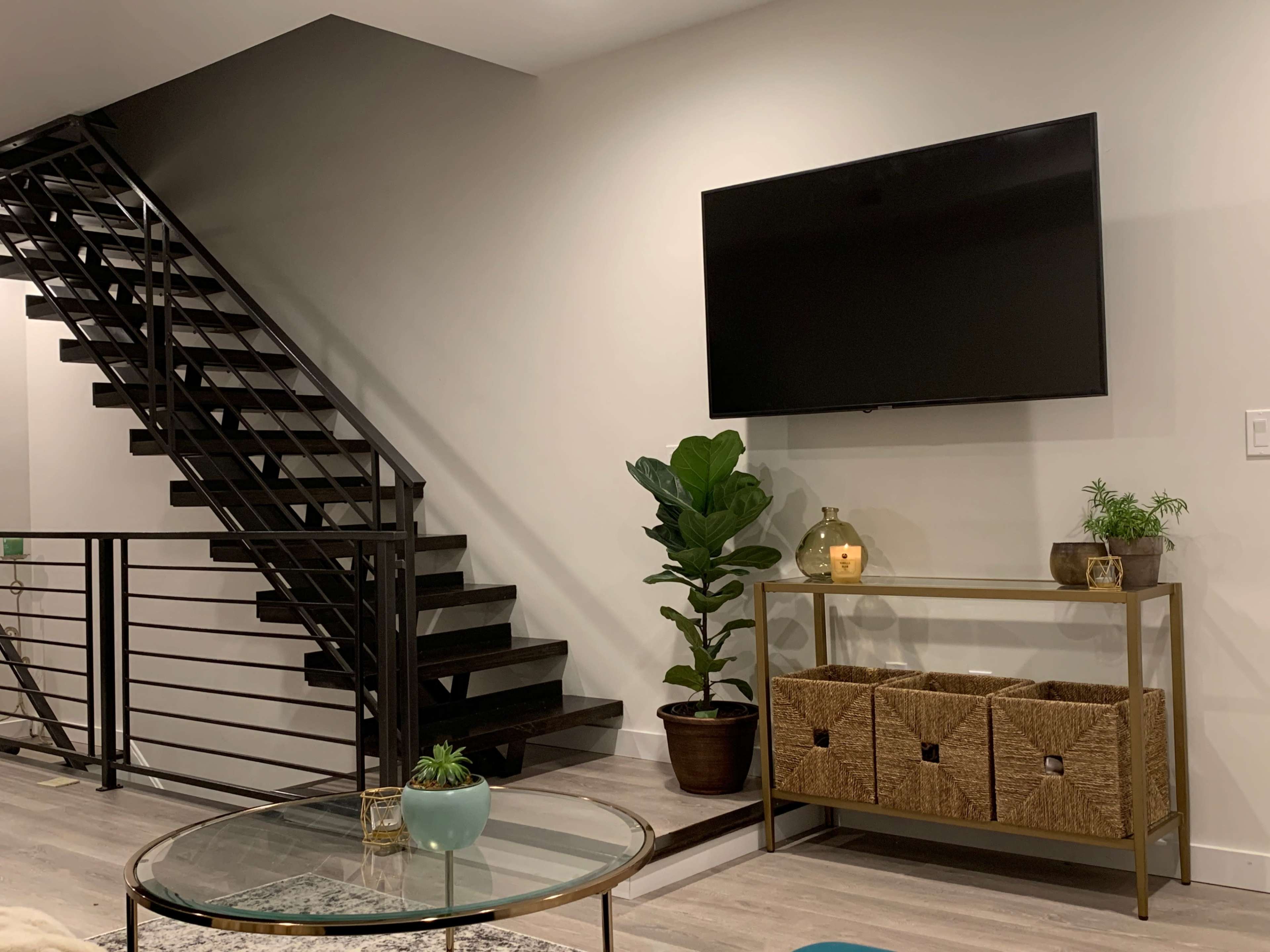 A modern living space features a staircase, a wall-mounted television, and a decorative shelf with storage baskets and plants.