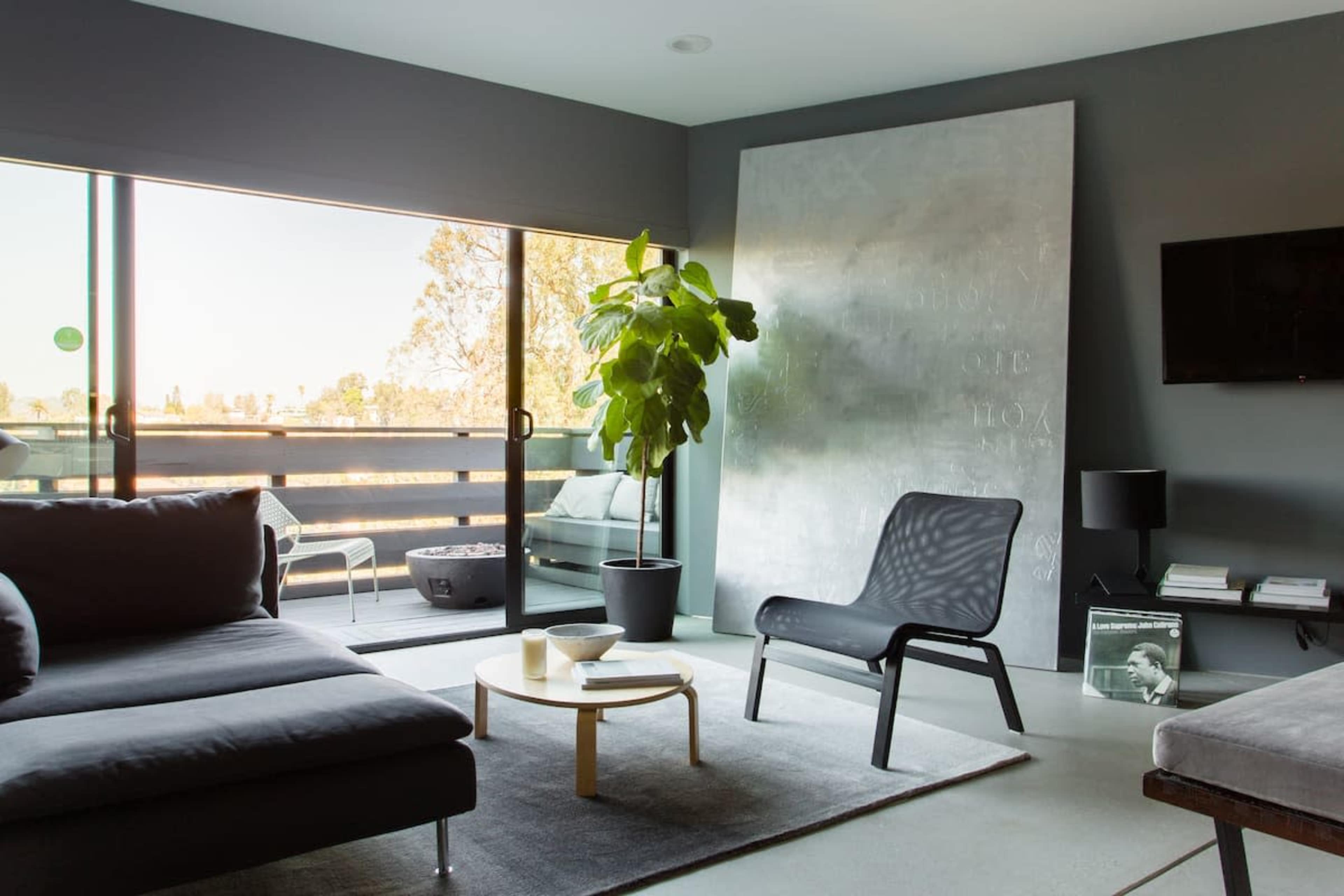 A modern living room features a gray sofa, a sleek chair, a round coffee table, and a large window overlooking an outdoor space with a plant beside it.