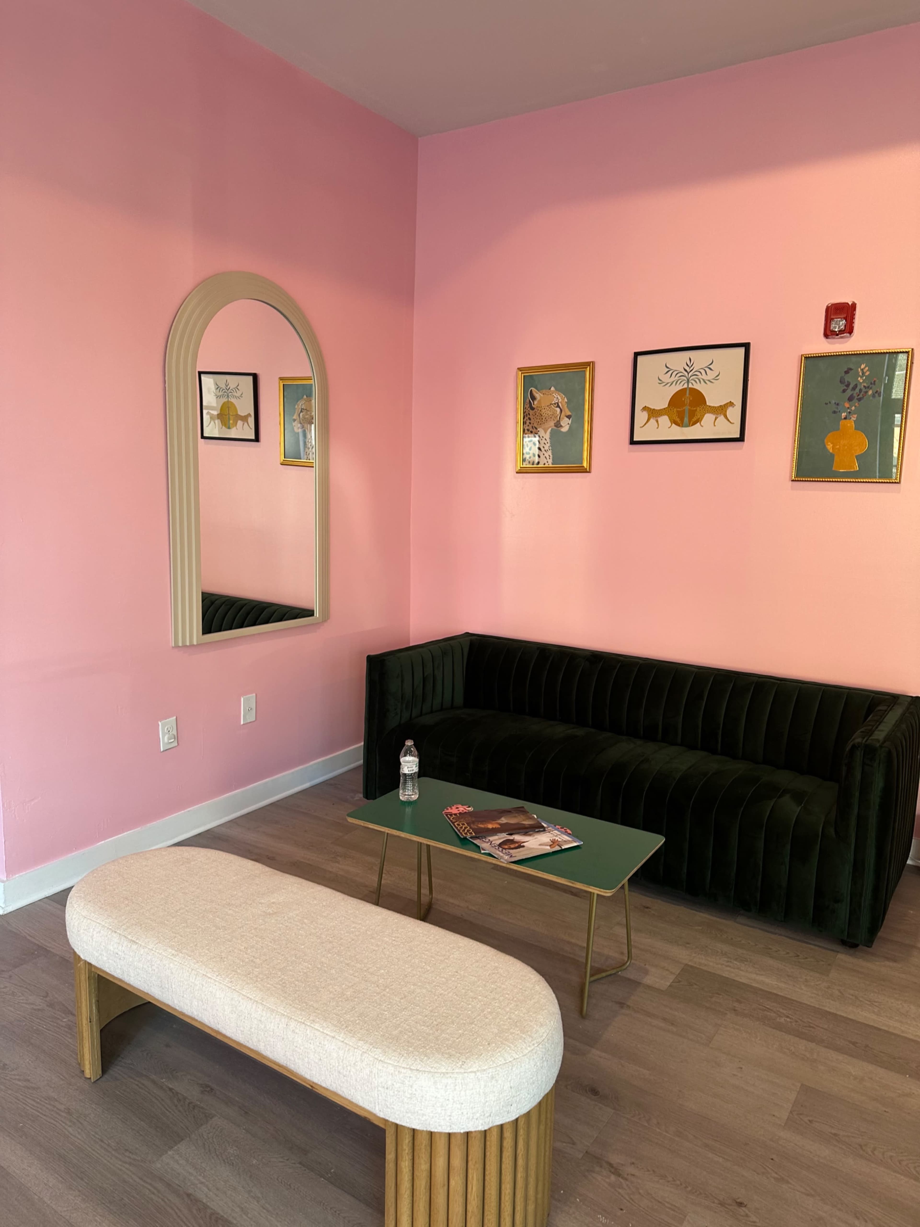 A room with pink walls featuring a green velvet couch, a light-colored bench, and a small table with magazines.