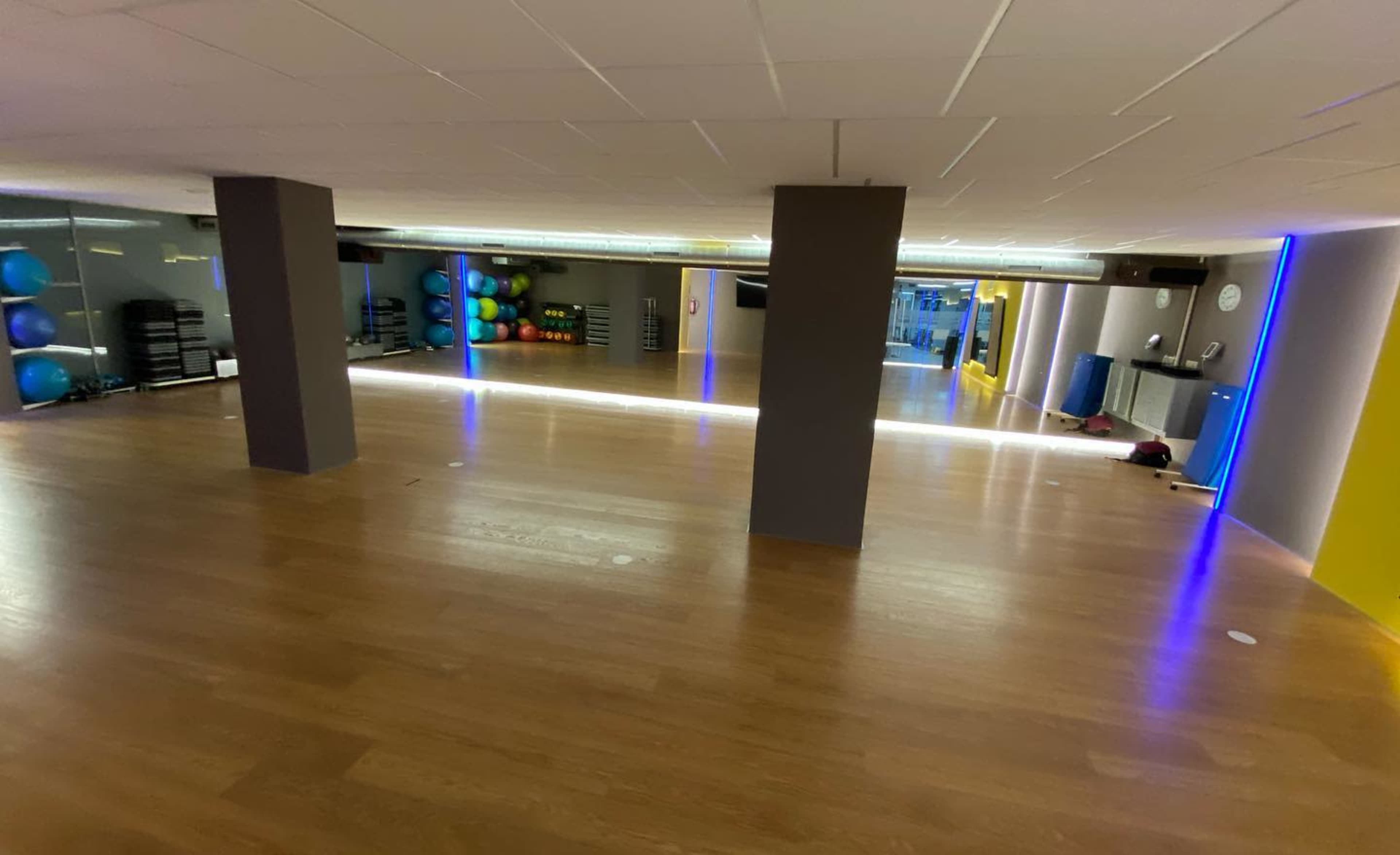 A spacious gym studio features wooden flooring, mirrored walls, and exercise equipment along the sides.