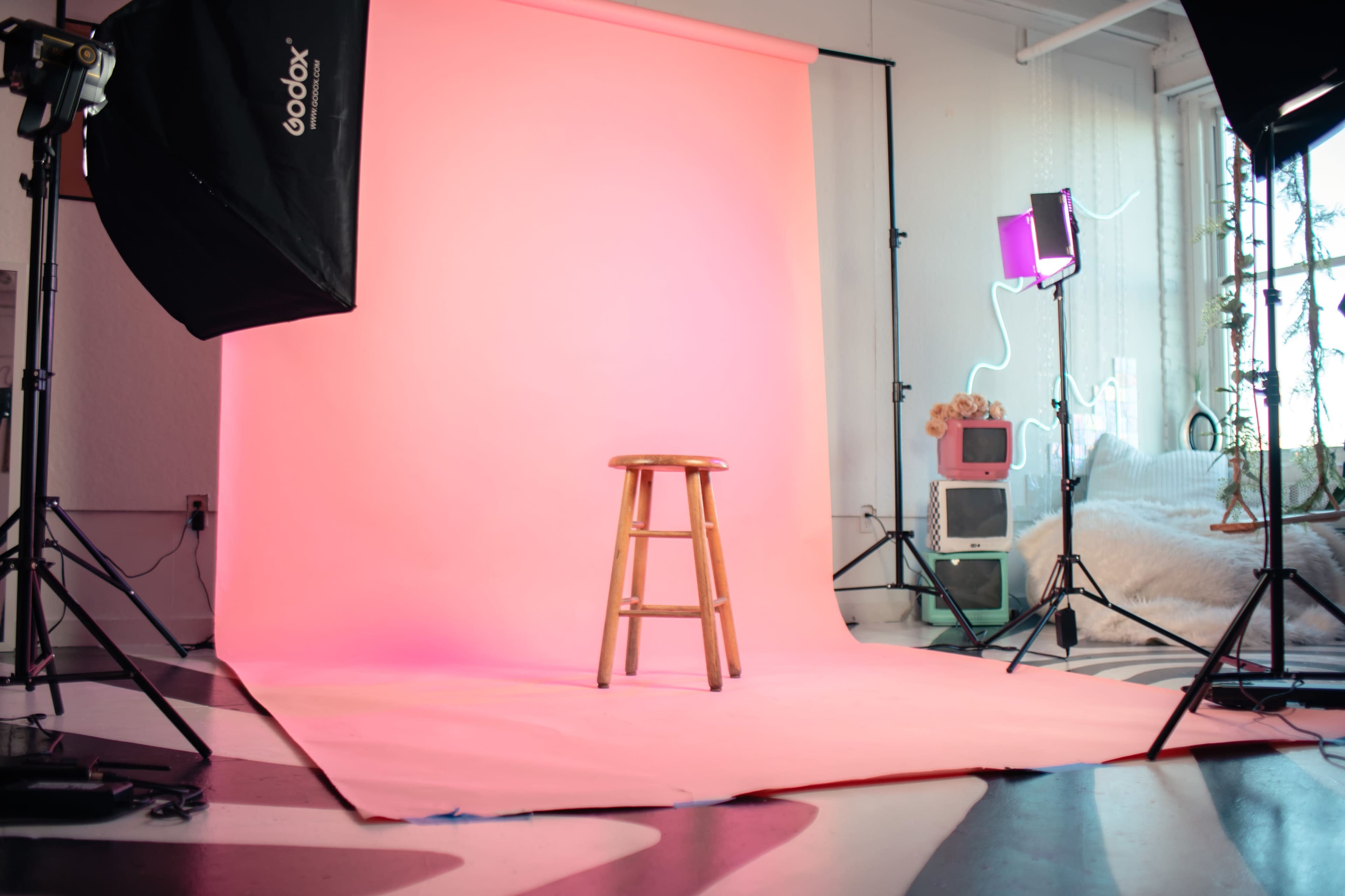 A photography studio setup featuring a pink backdrop, a wooden stool, and various lighting equipment arranged around the scene.