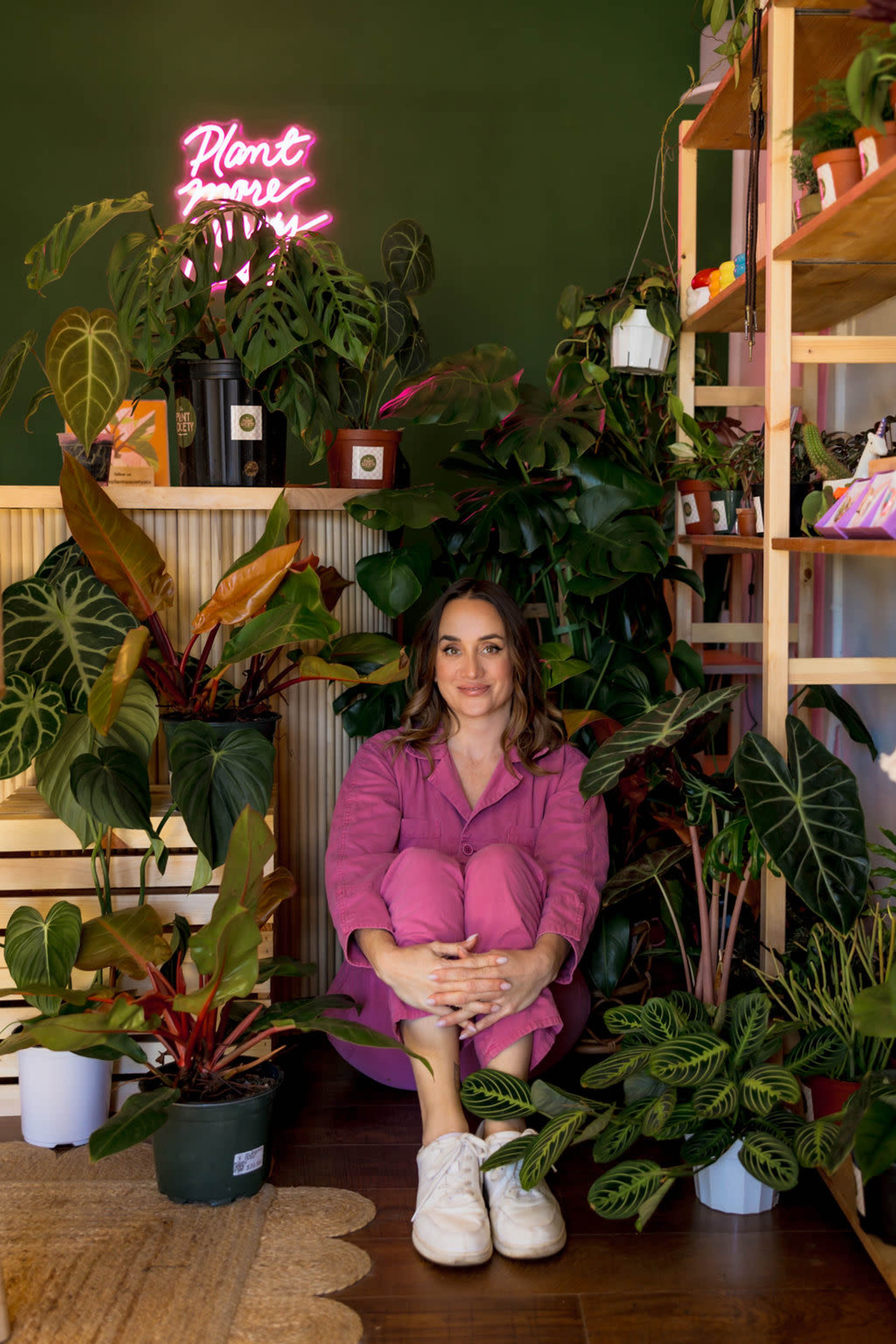 A woman sits cross-legged among various potted plants, with a neon sign in the background reading "Plant More."