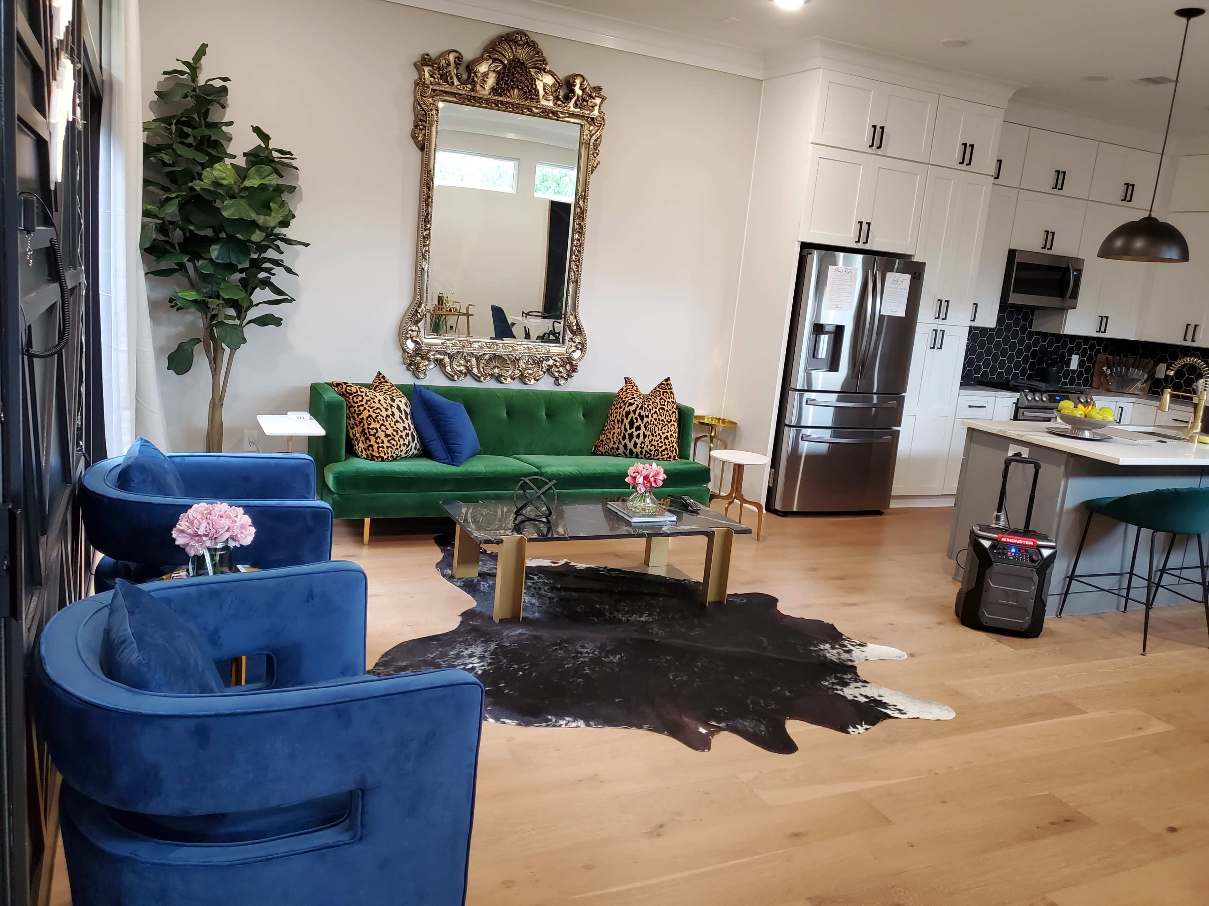 A modern living area featuring a green velvet sofa with patterned pillows, two blue chairs, a glass coffee table, and a kitchen area in the background.