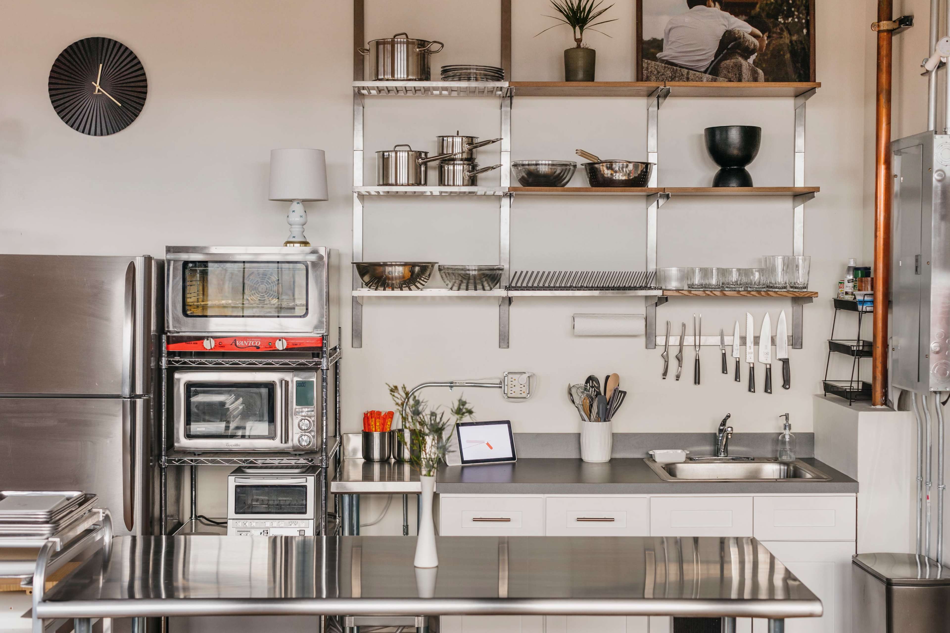 A modern kitchen with metal shelves holding cookware, a countertop with a sink, and various kitchen appliances, including microwaves and a clock on the wall.