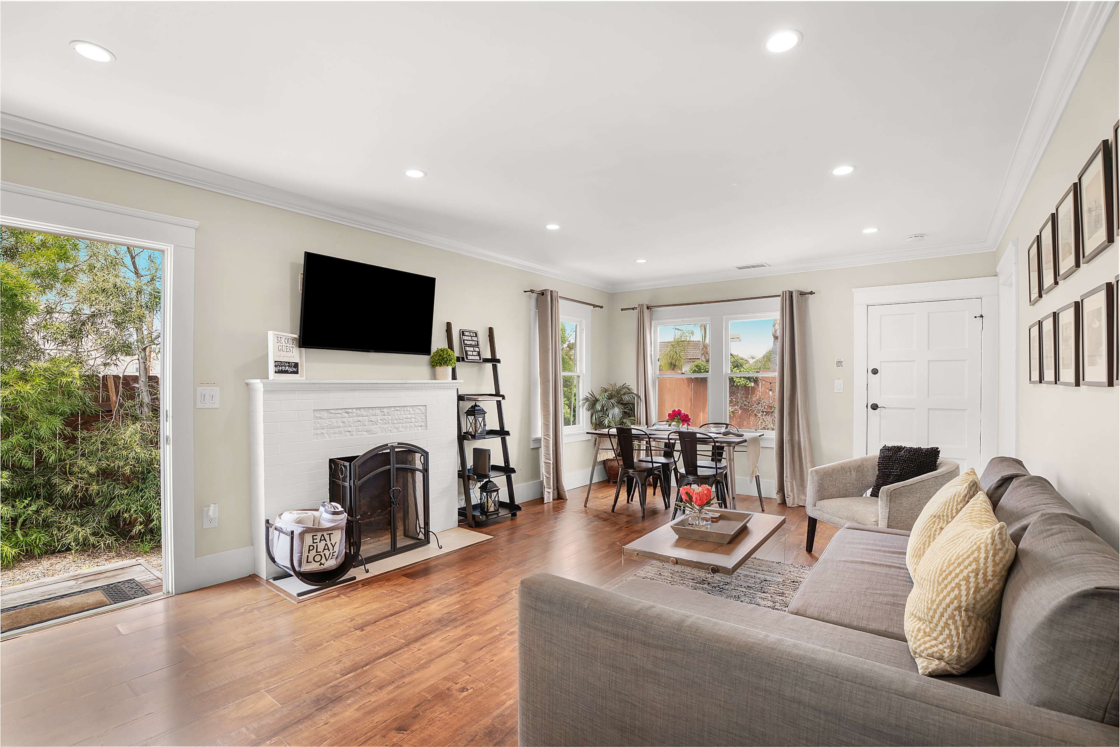 The living room features a gray sofa, a fireplace, a television mounted on the wall, and a dining area with a table set for four, alongside large windows and an entrance to the outside.