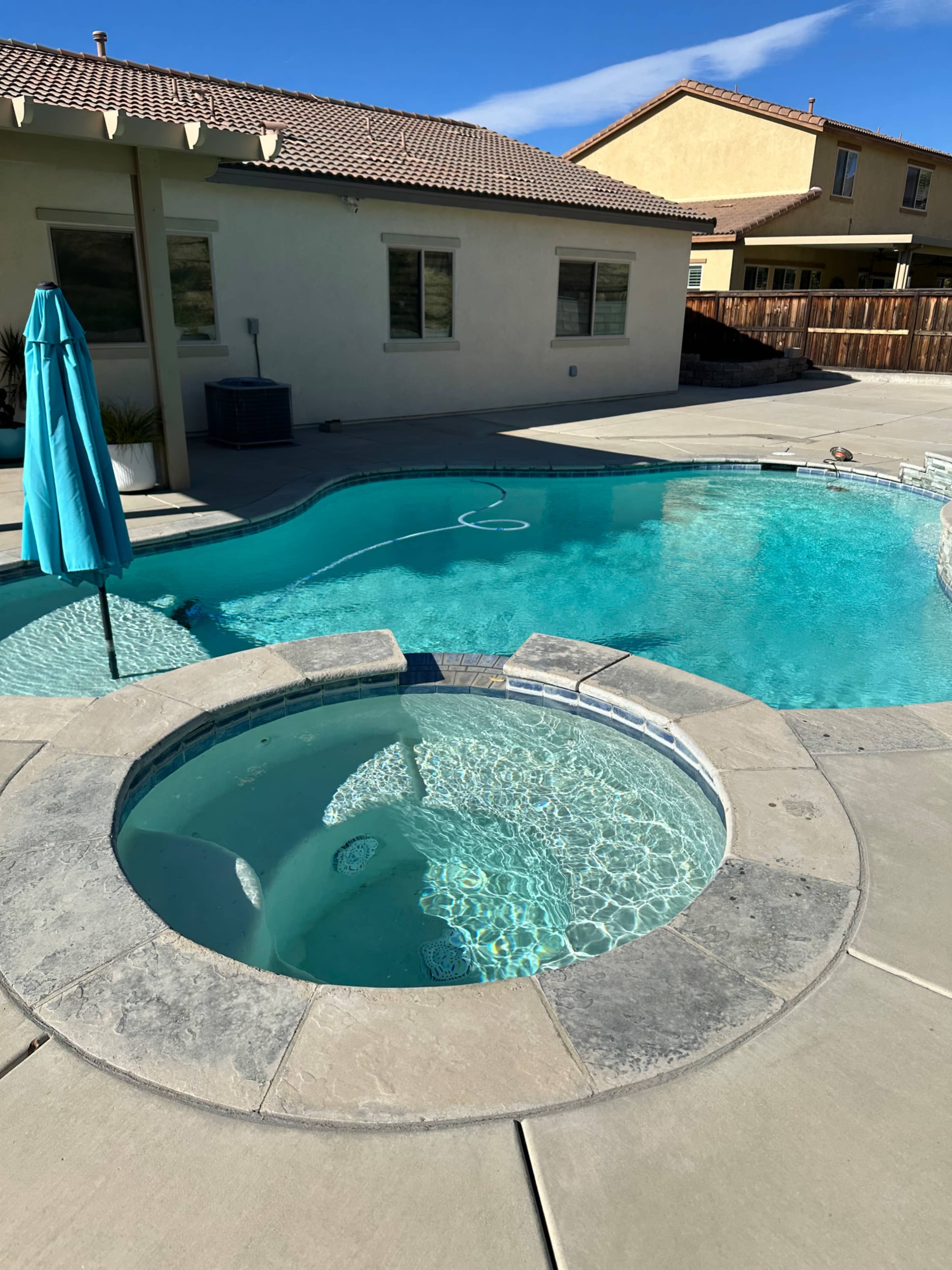 The image shows a swimming pool with an attached hot tub in a residential backyard, featuring a tall blue umbrella nearby and a clear blue sky above.