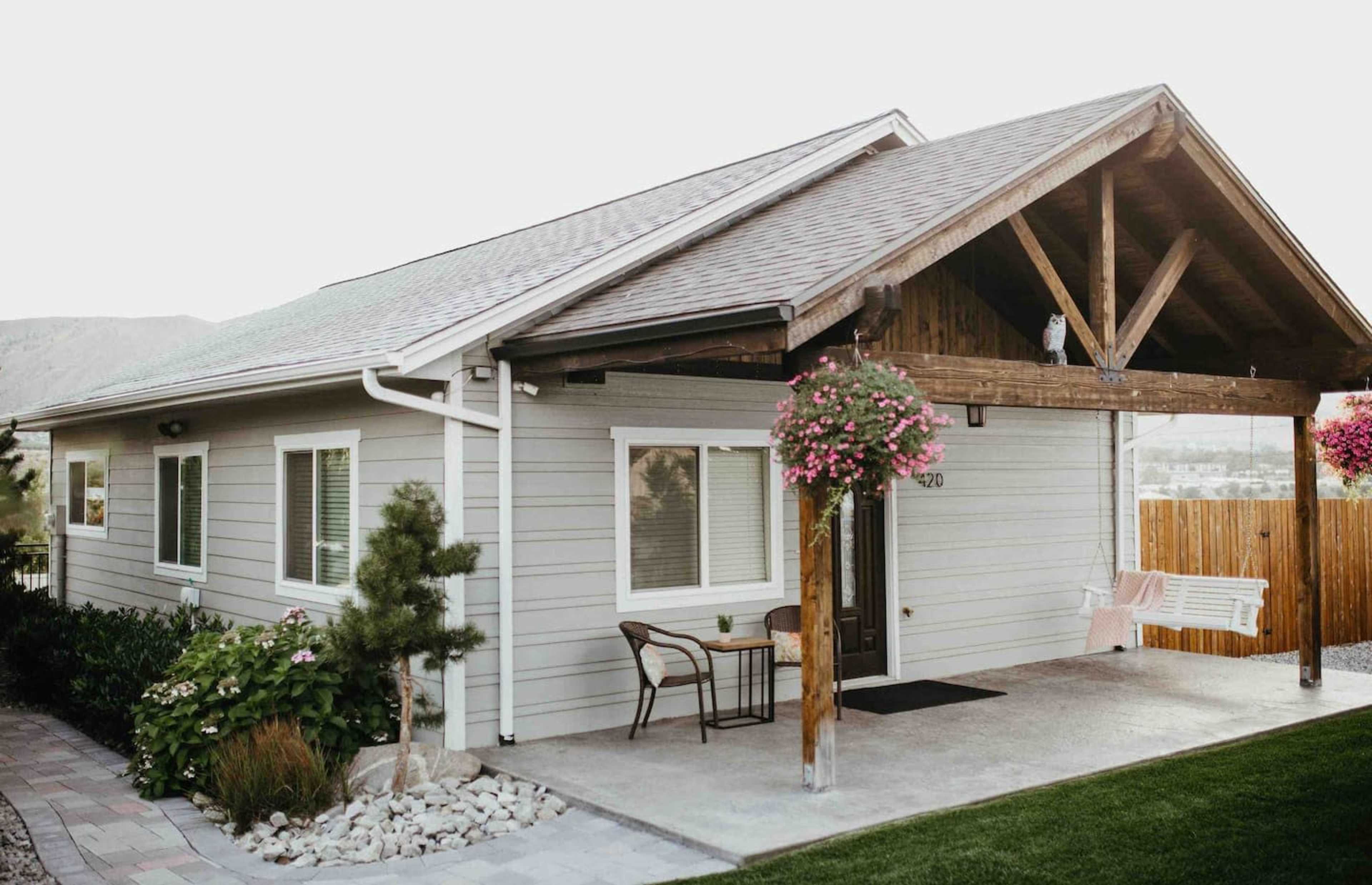 A single-story house with a gabled roof features a front porch supported by wooden beams, surrounded by landscaped greenery and hanging flower baskets.