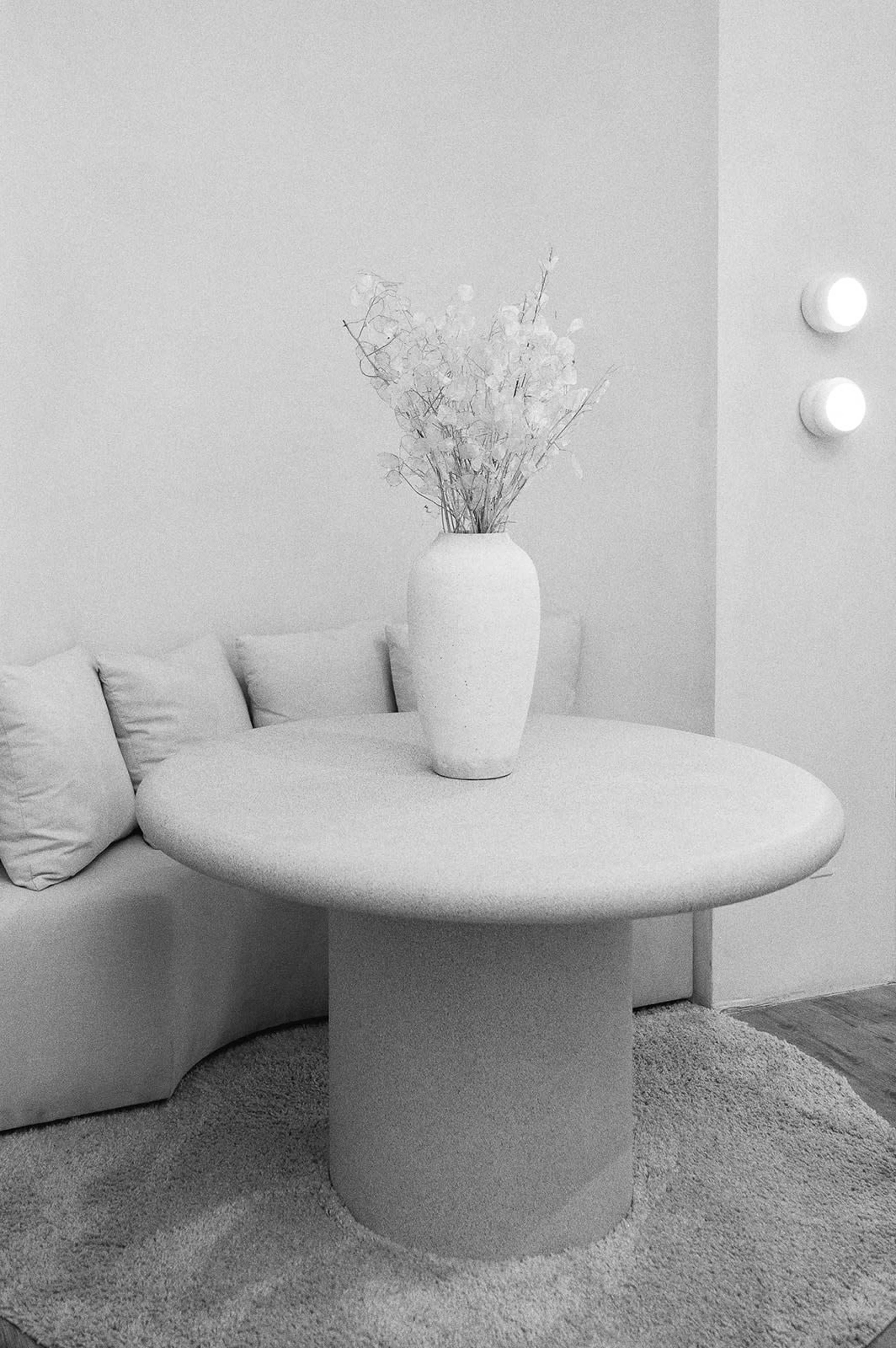 A round table with a textured surface sits on a rug, topped by a minimalist vase holding decorative branches, in a softly lit room with a neutral color palette.