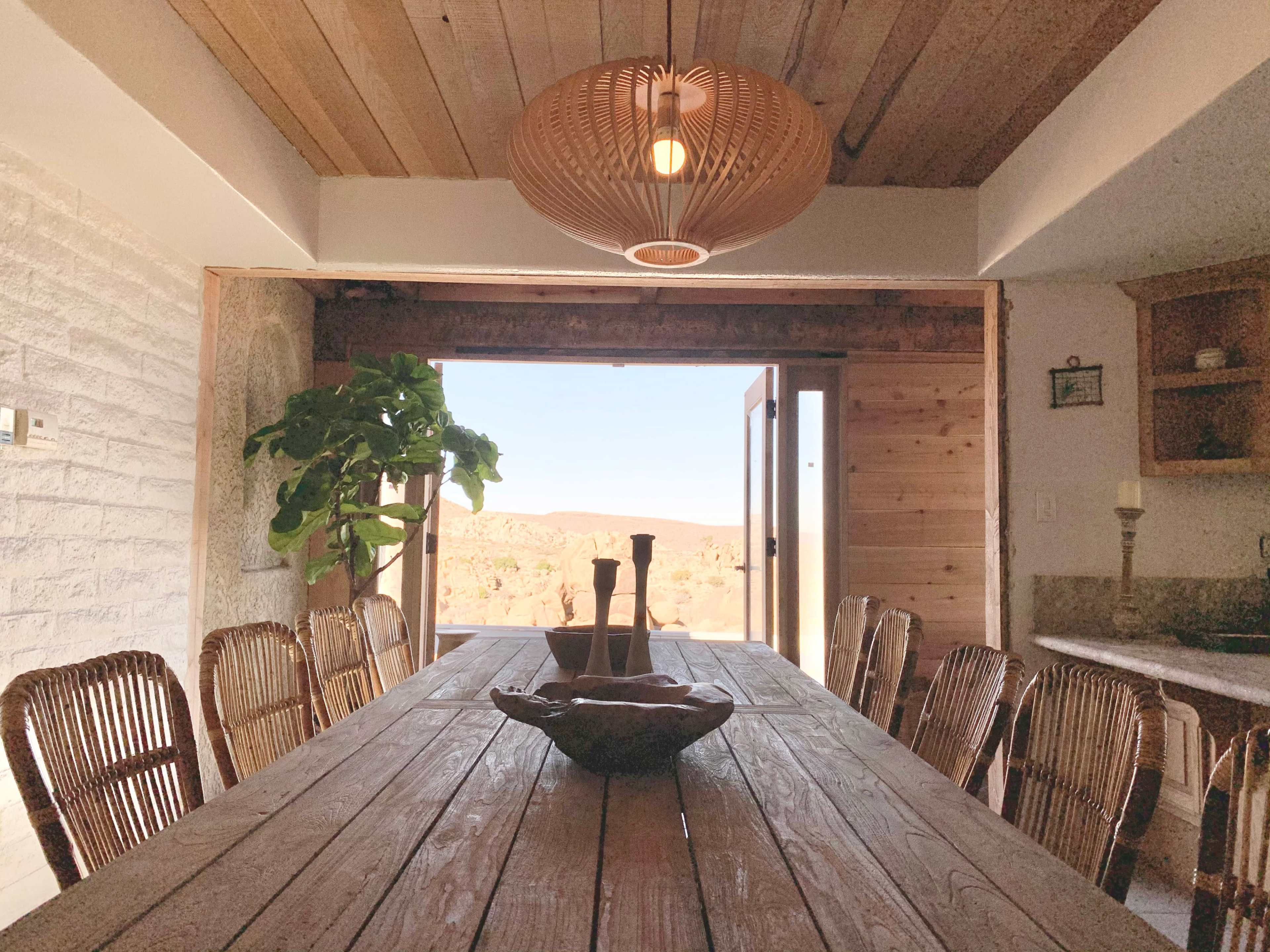 A wooden dining table with woven chairs is set in a room with a large open door revealing a view of a desert landscape.