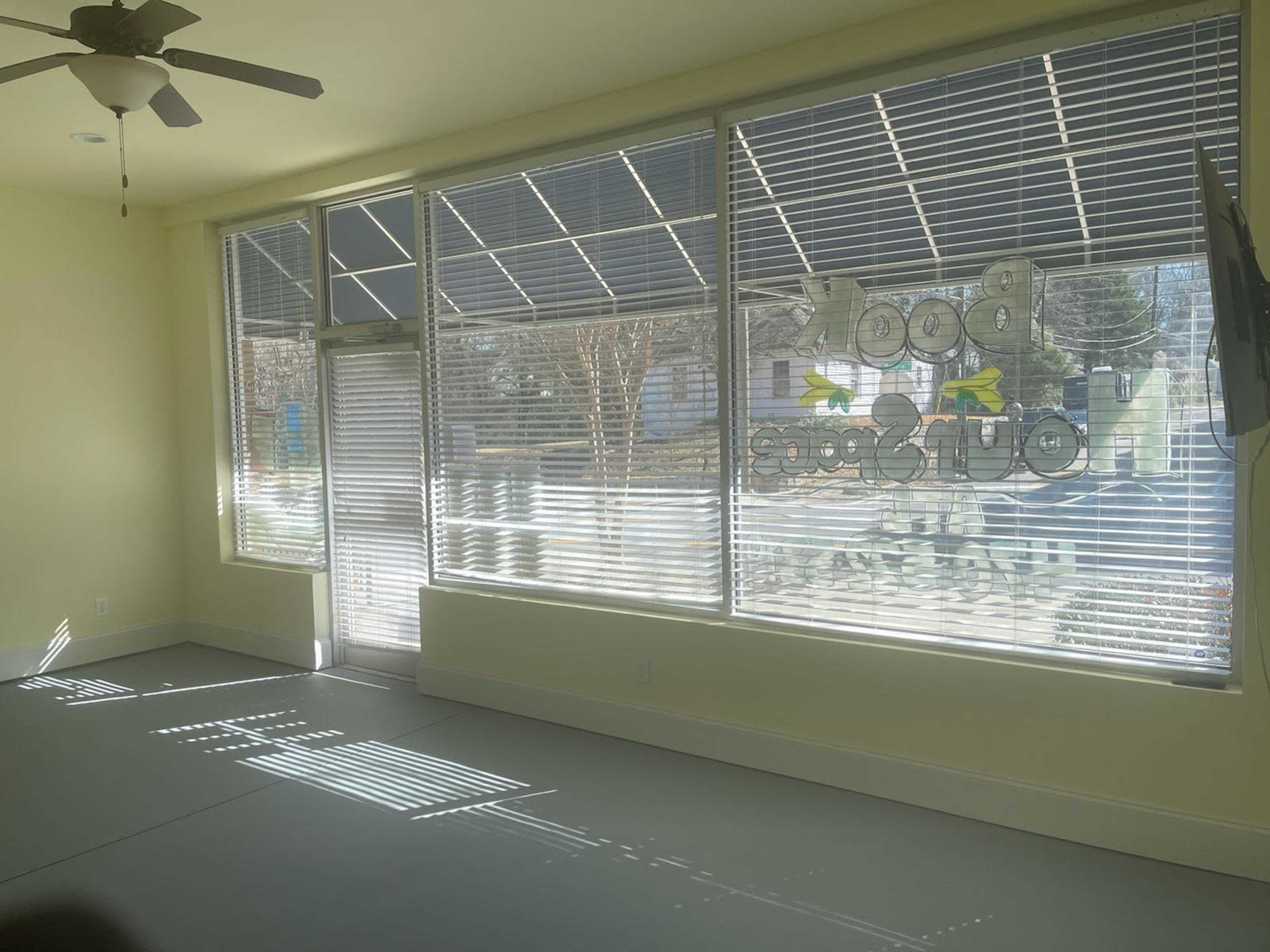A brightly lit room features large windows with blinds, casting shadows on a light-colored floor, and a door leading outside.