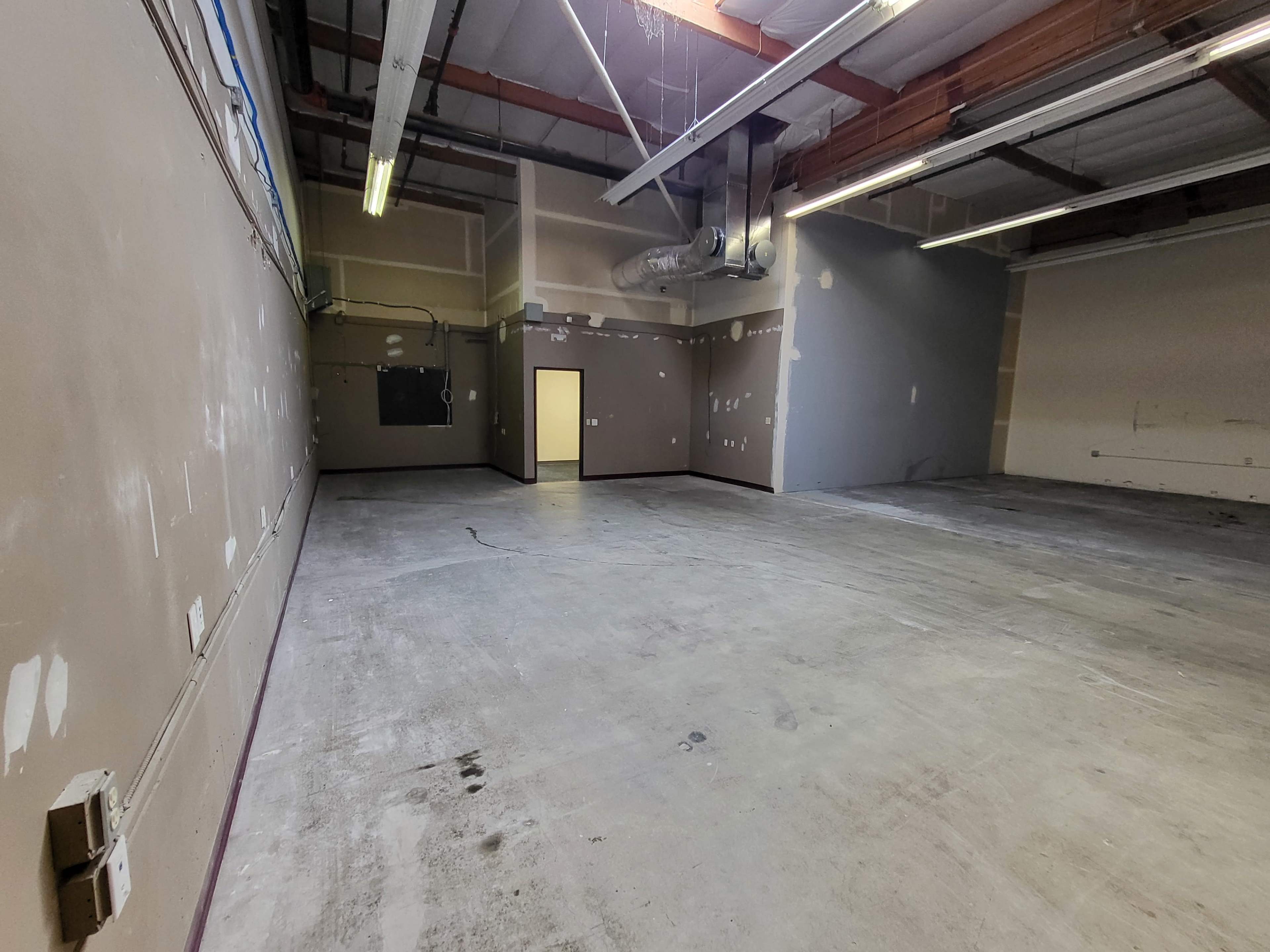 The image shows an empty industrial space with concrete flooring and gray walls, featuring a single door and overhead lighting.