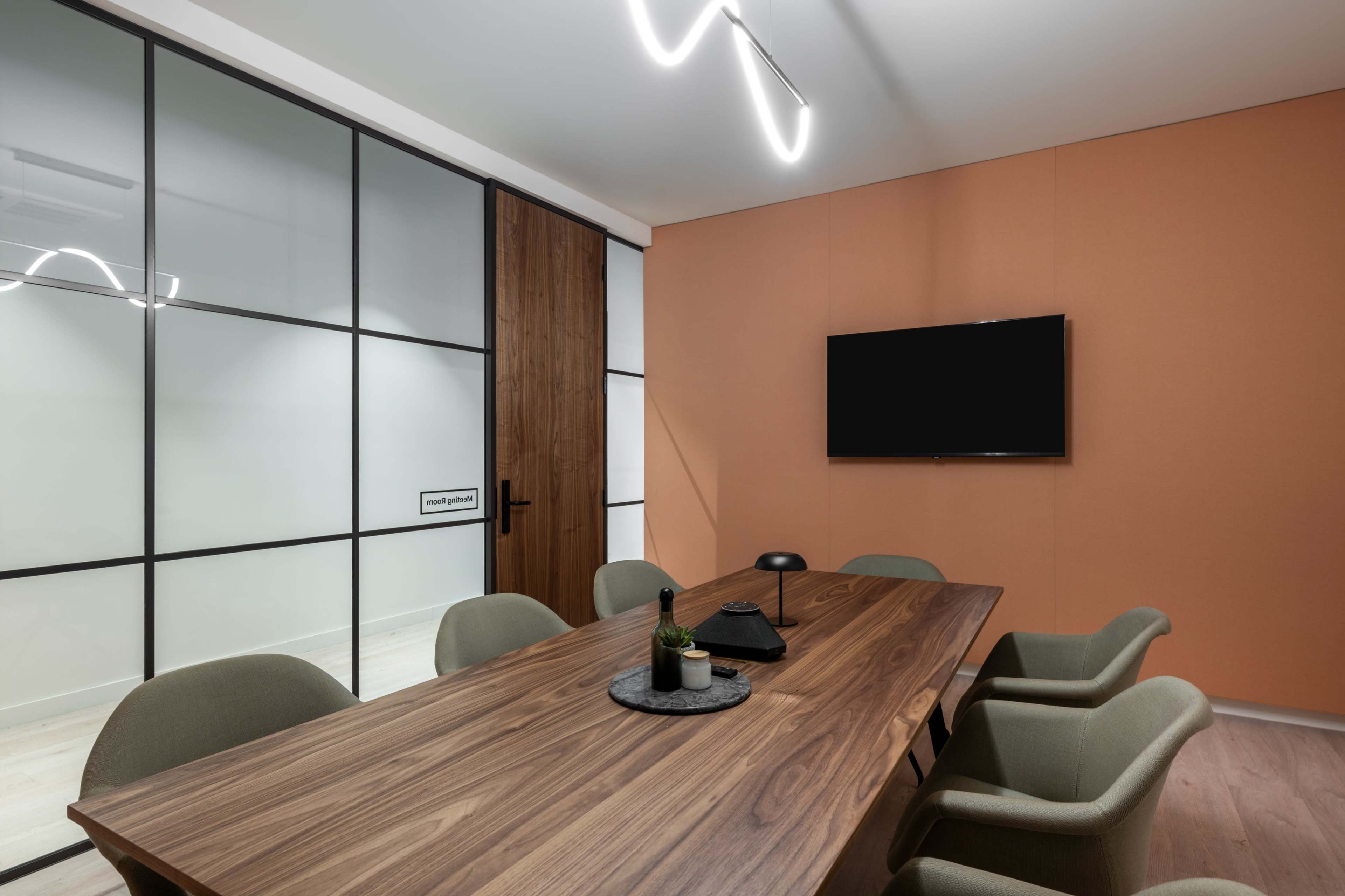 A conference room features a wooden table surrounded by green chairs, with a wall-mounted television and glass partition dividing the space.