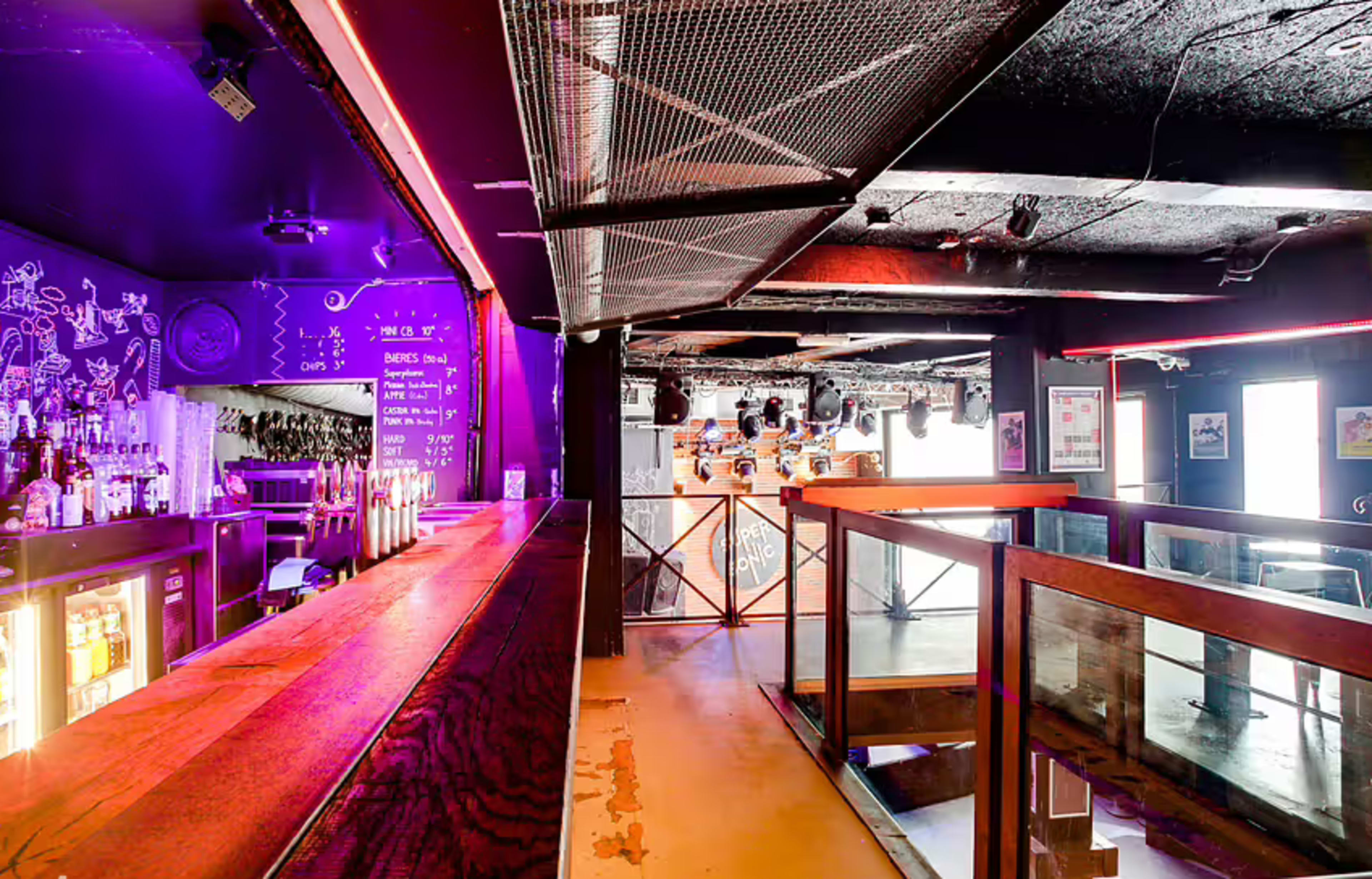 The image shows a dimly lit venue with a long bar, illuminated by pink and purple lights, and a stage area visible in the background.