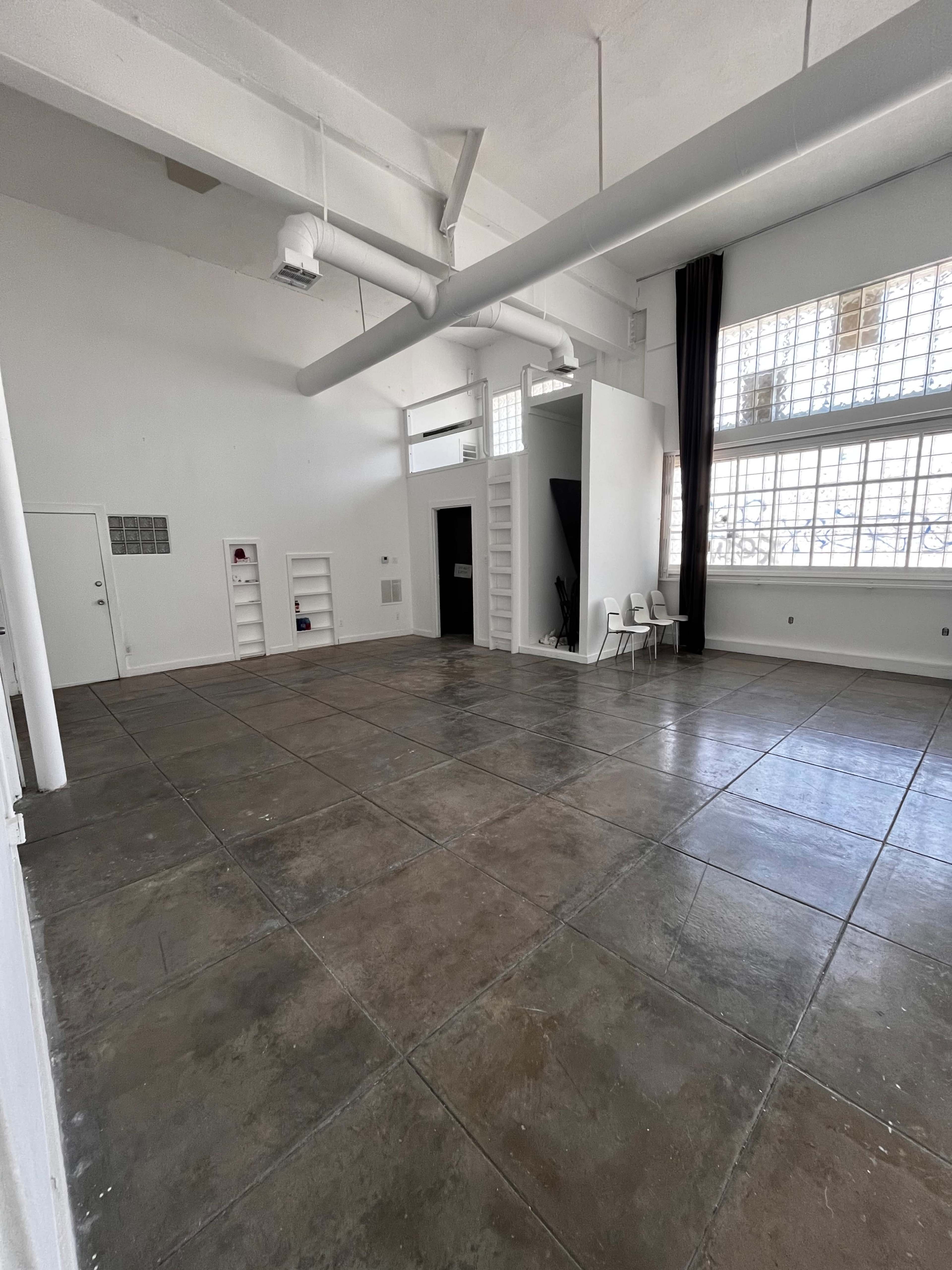 The image shows a spacious, empty studio with polished concrete floors, large windows, and minimal furnishings.