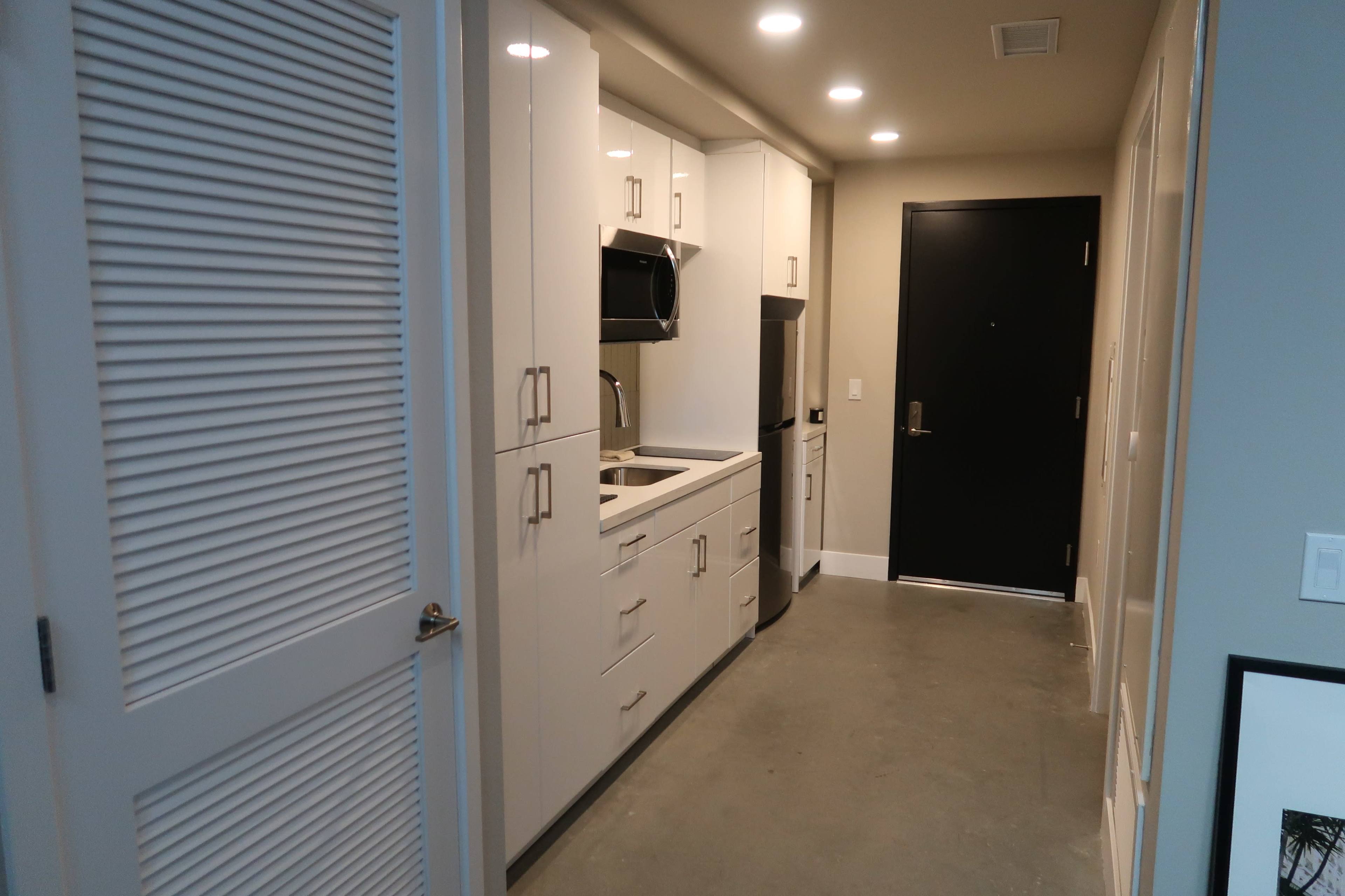 The image shows a narrow hallway leading to a kitchen area with white cabinets, an open door, and concrete flooring.