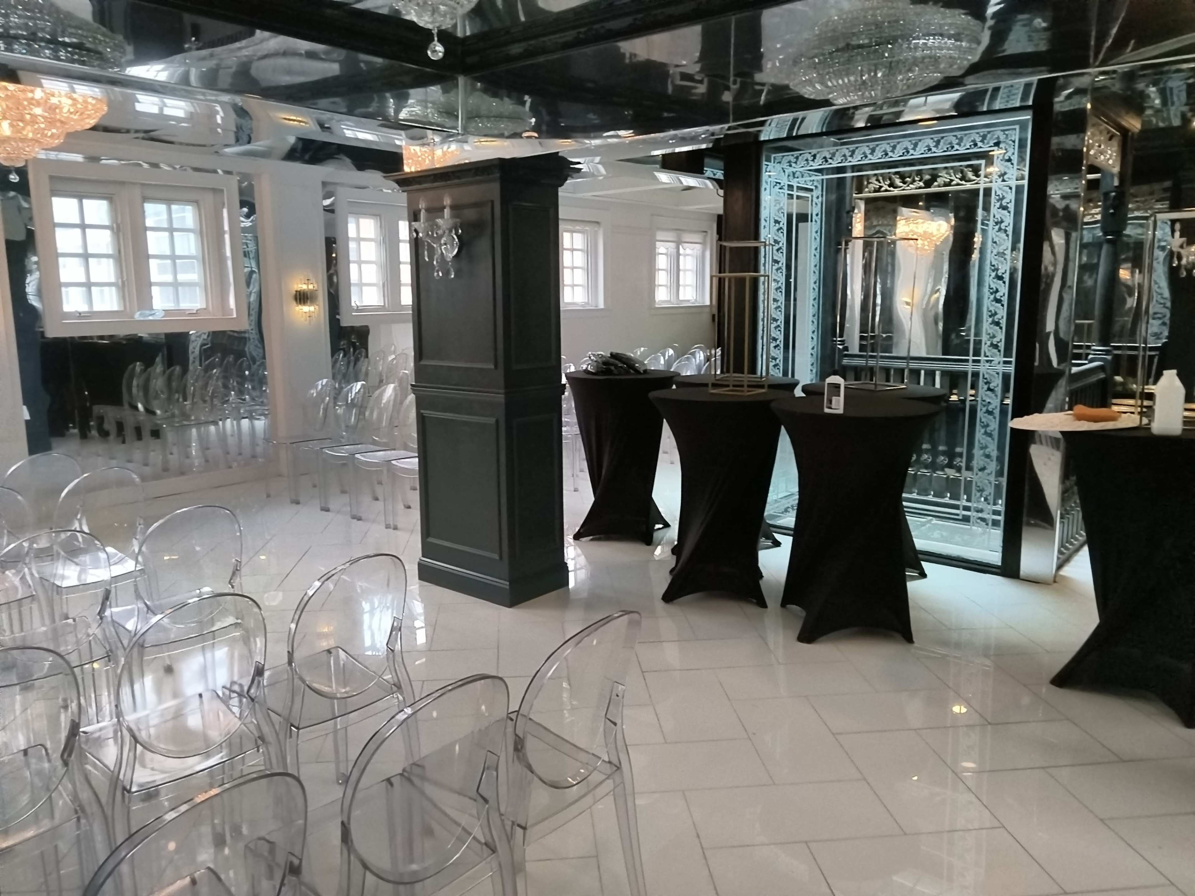 The image shows a modern event space with transparent chairs arranged in rows and two black cocktail tables near a mirrored wall.