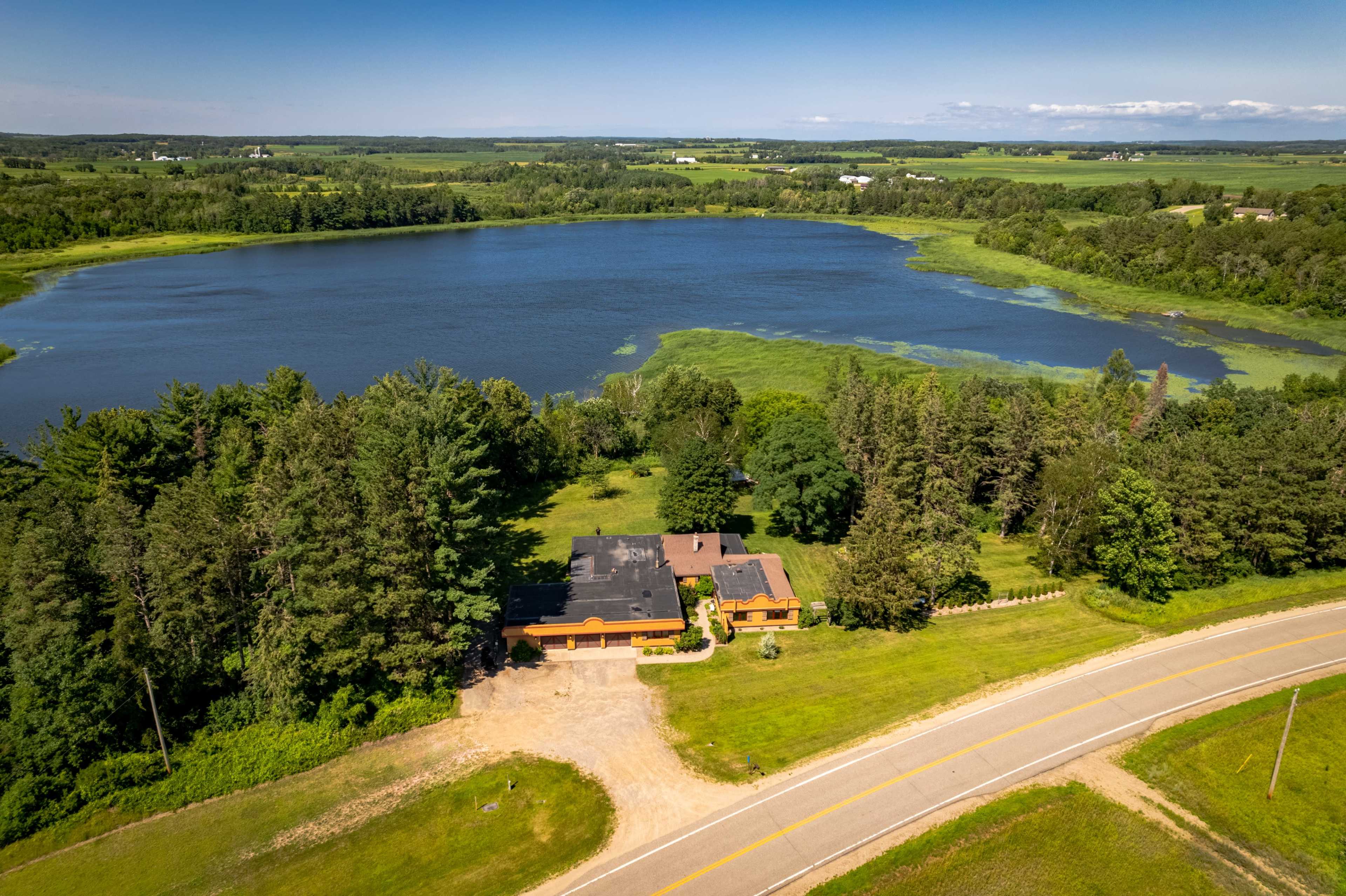 A single-story house is situated near a large lake, surrounded by trees and greenery, with a road running alongside it.